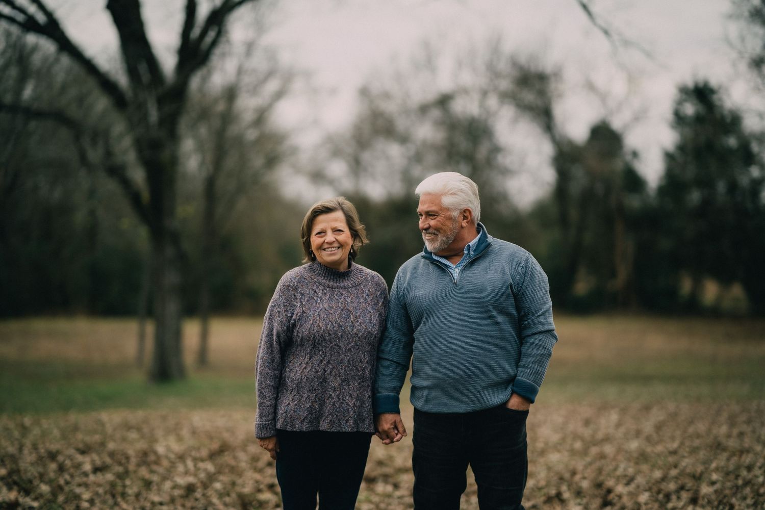 A couple shares a tender moment in an autumn field surrounded by bare trees.