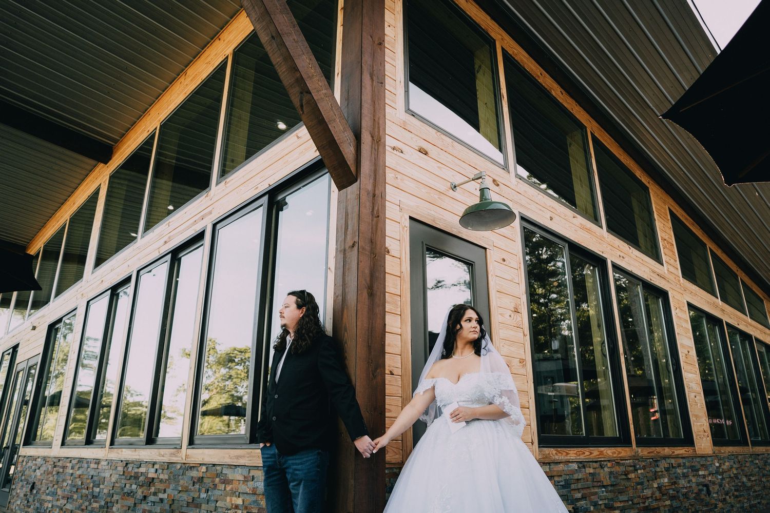 A couple stands outside a modern glass and wood building in wedding attire.