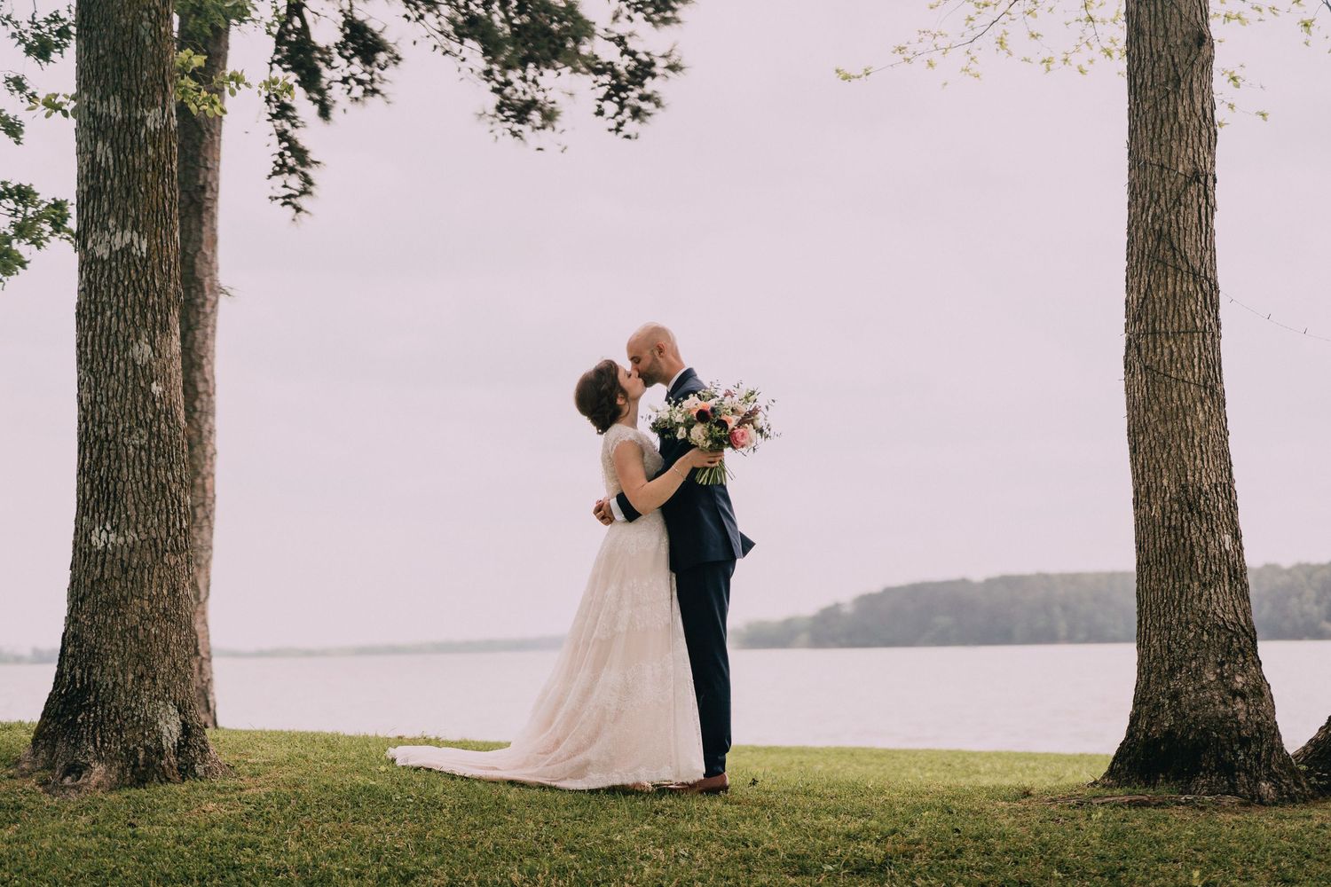 A romantic wedding couple embraces between two tall trees overlooking a serene waterfront view.