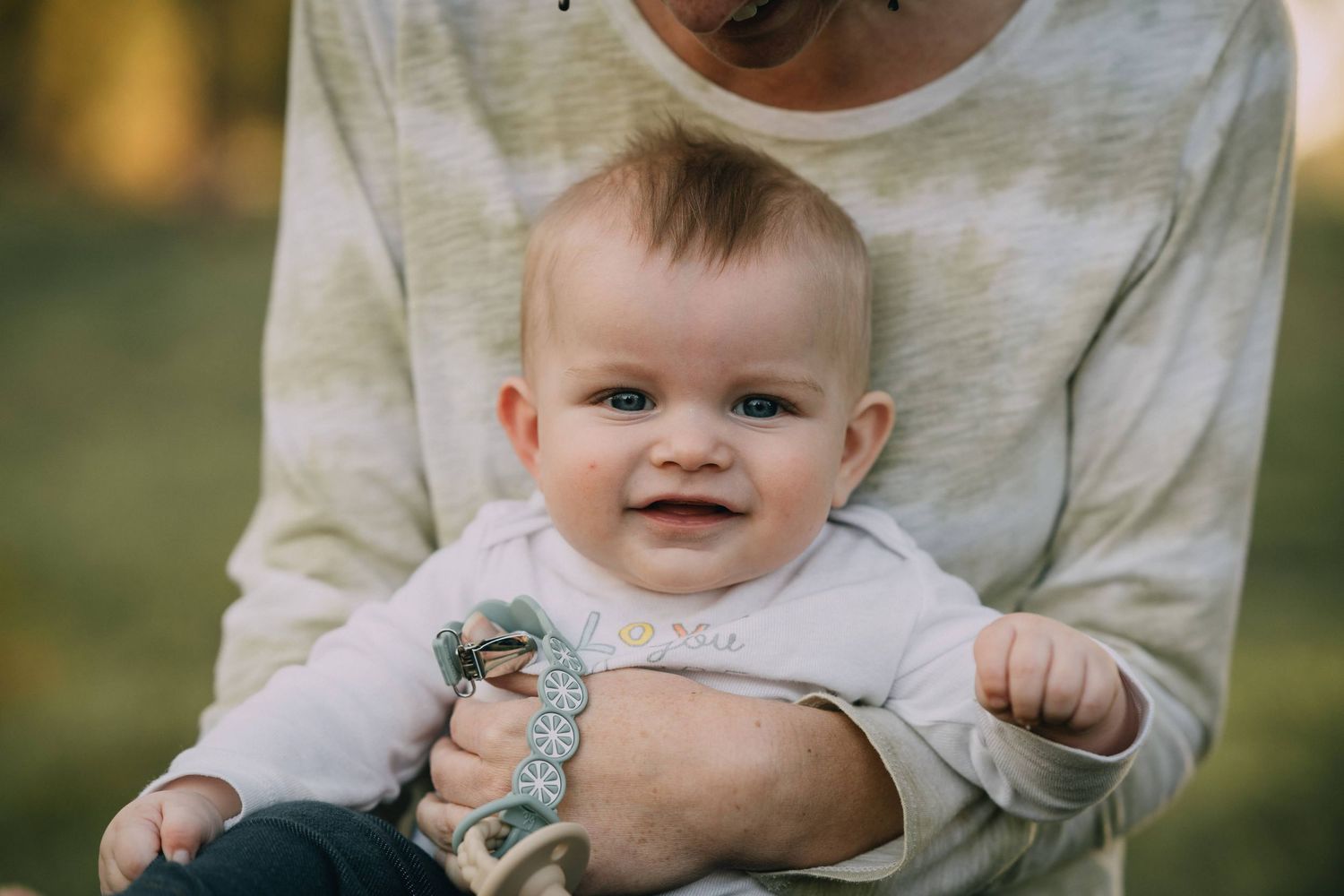 Baby wearing white clothing sitting on someone's lap outdoors.