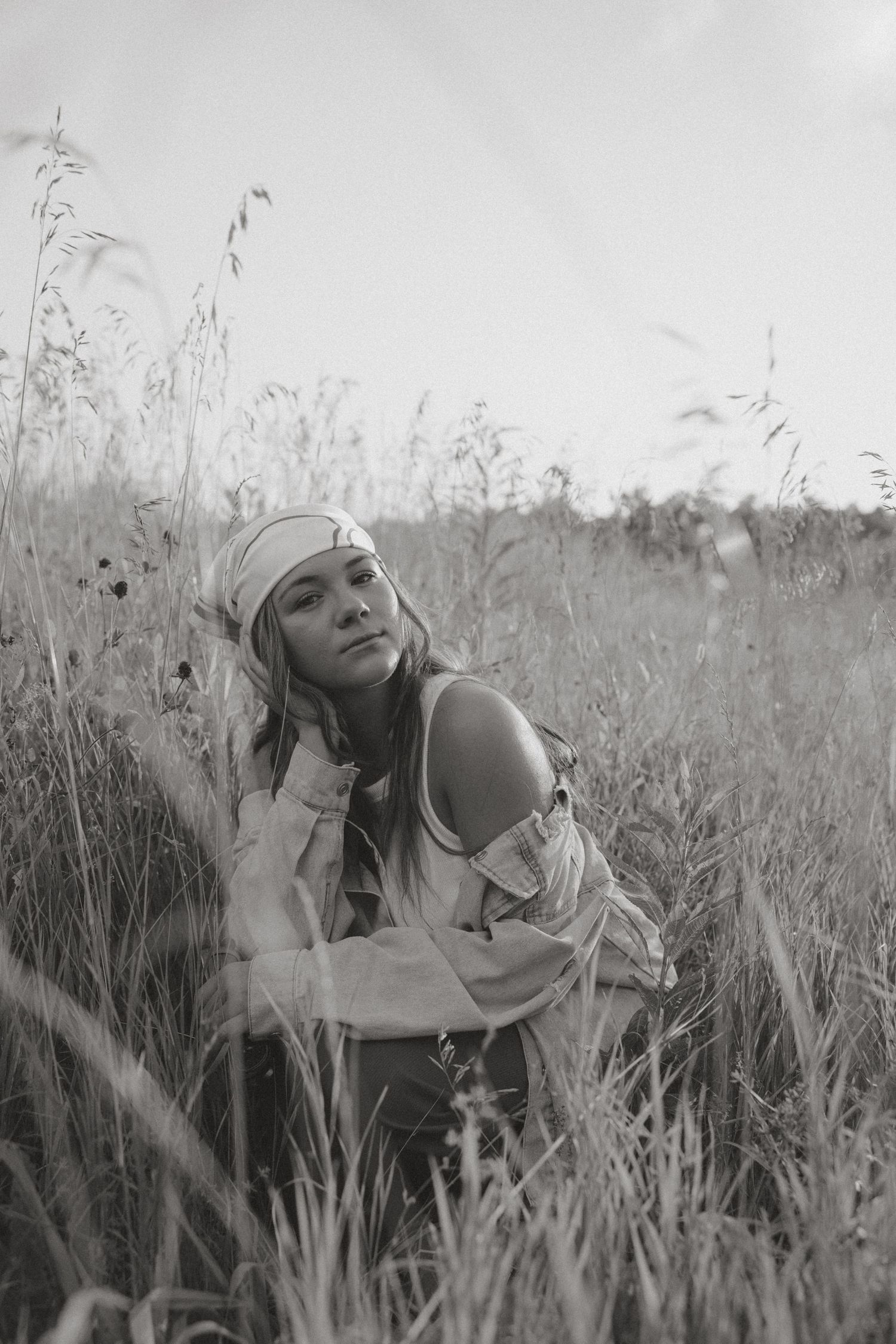 Black and white portrait of a woman sitting in tall grass field looking upward.