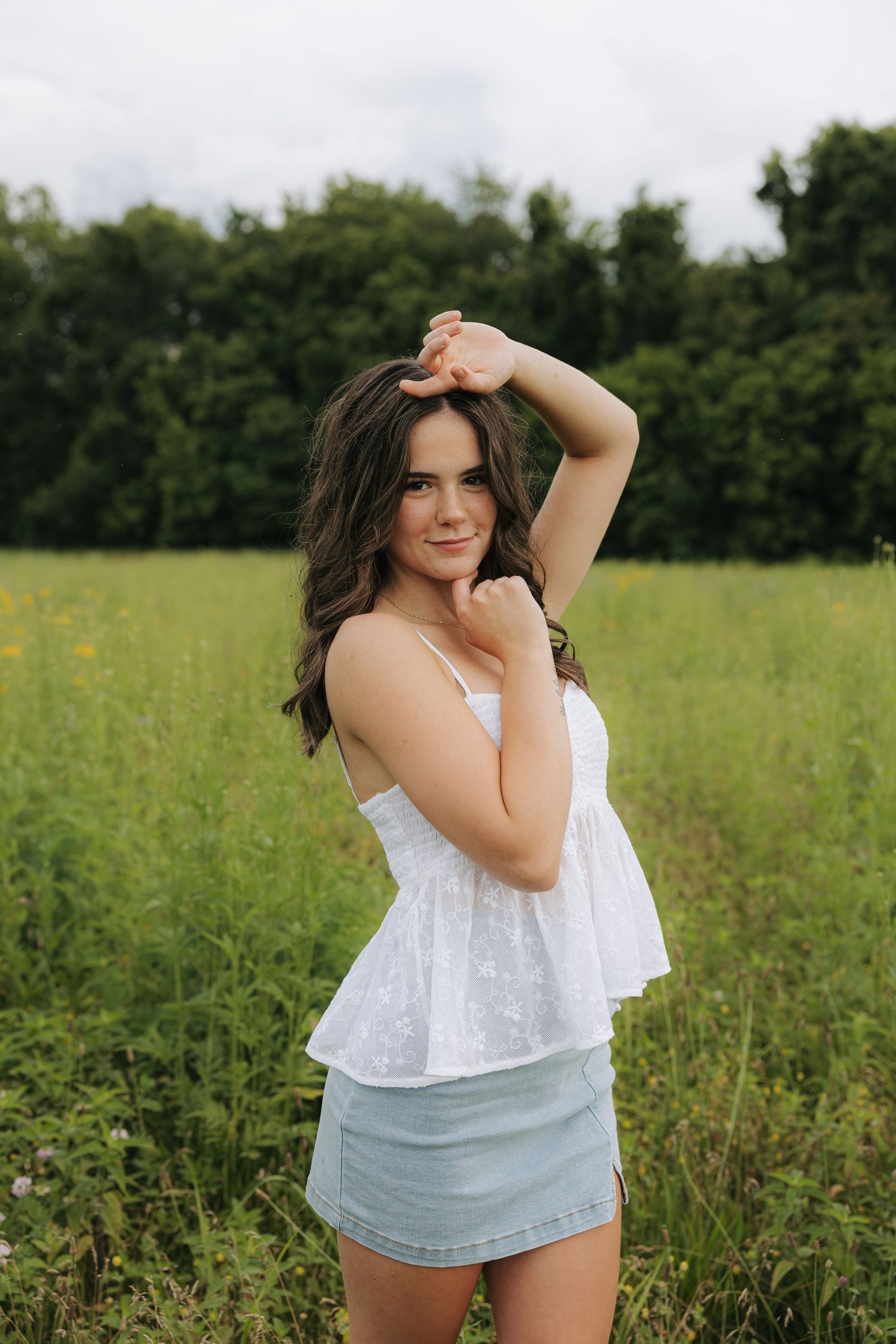 Young brunette woman in white top and denim skirt posing in a green meadow.
