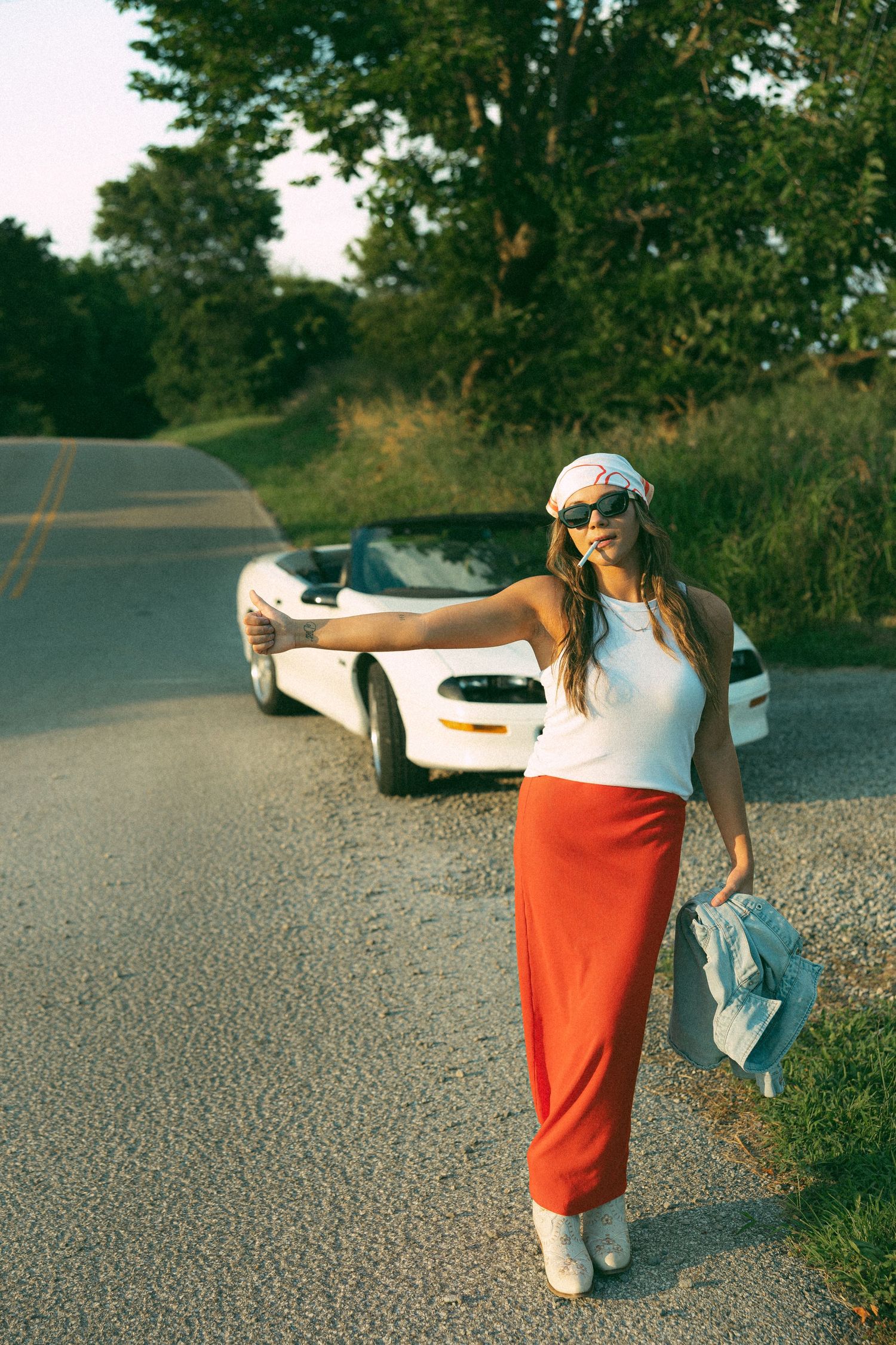 Woman in red skirt and white top hitchhiking on a rural road beside a white convertible at sunset.