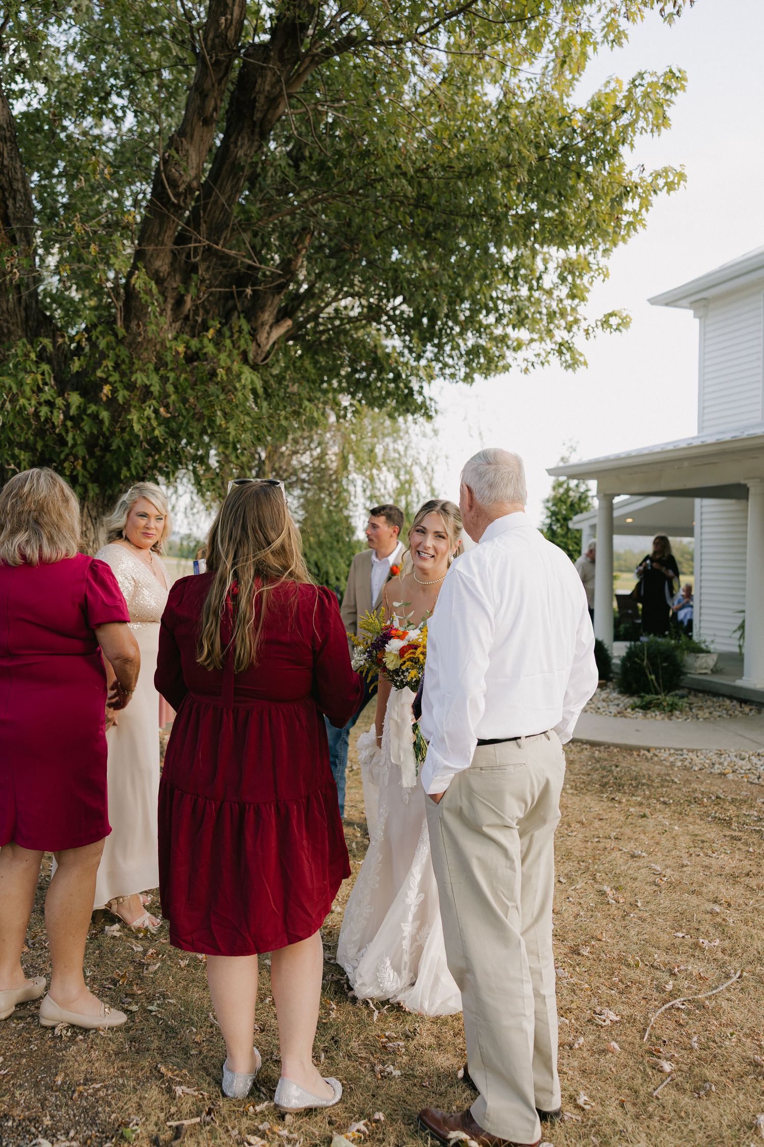 Bride holding colorful bouquet surrounded by family members conversing outdoors near a white building.