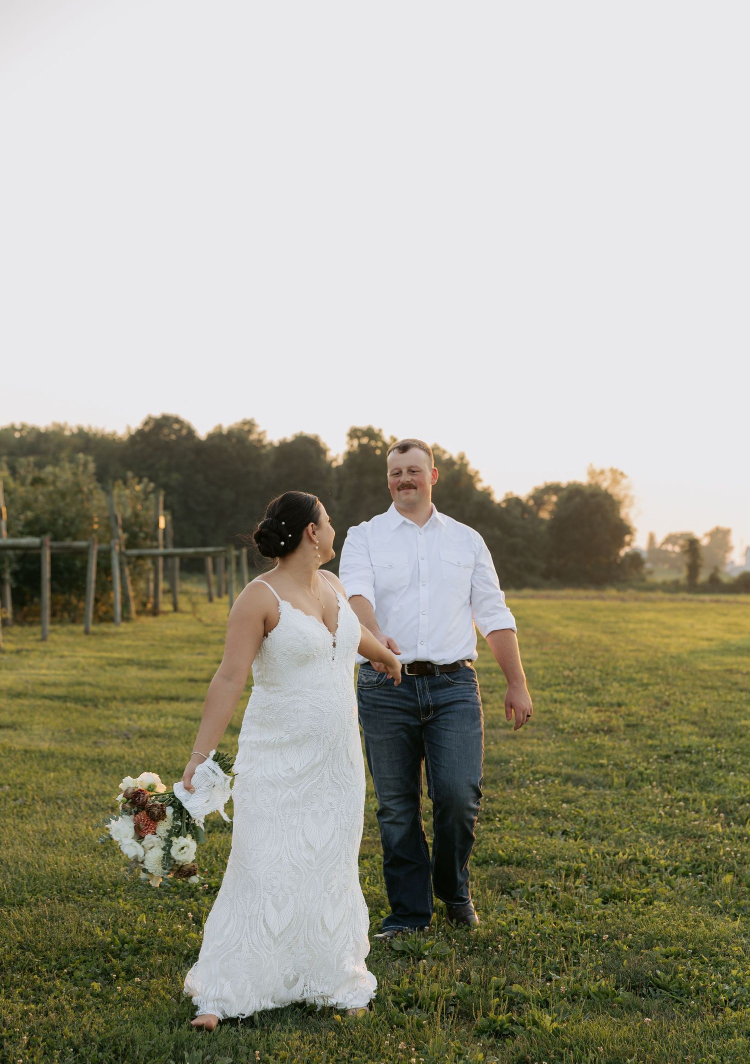 Bride and groom walking hand in hand through a green field at golden hour.