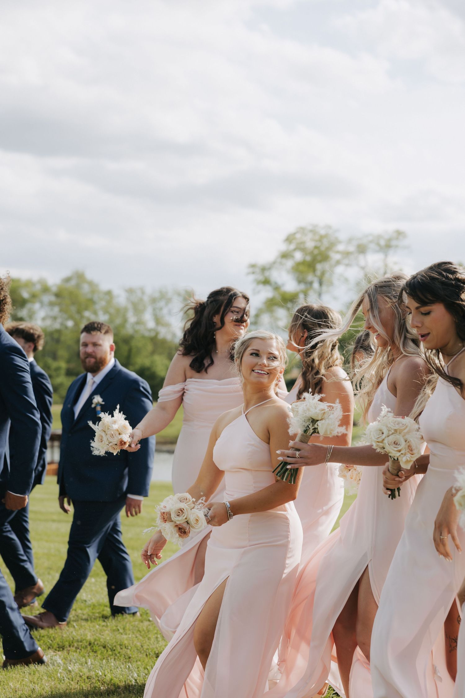 Bridesmaids in blush pink gowns carrying white bouquets walking with groomsmen at a wedding.