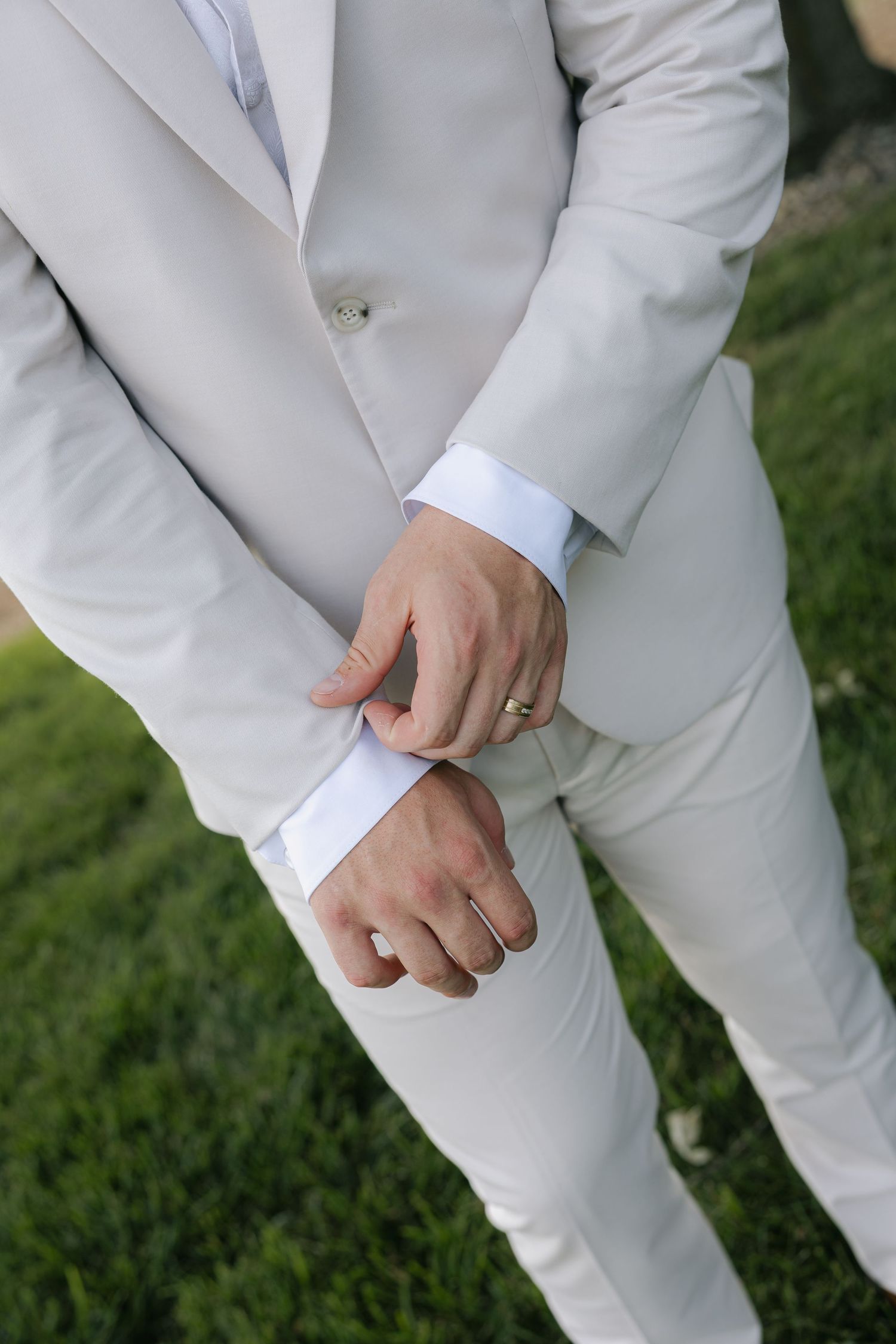 Close-up of groom in cream suit adjusting cufflinks with wedding ring visible on finger outdoors.