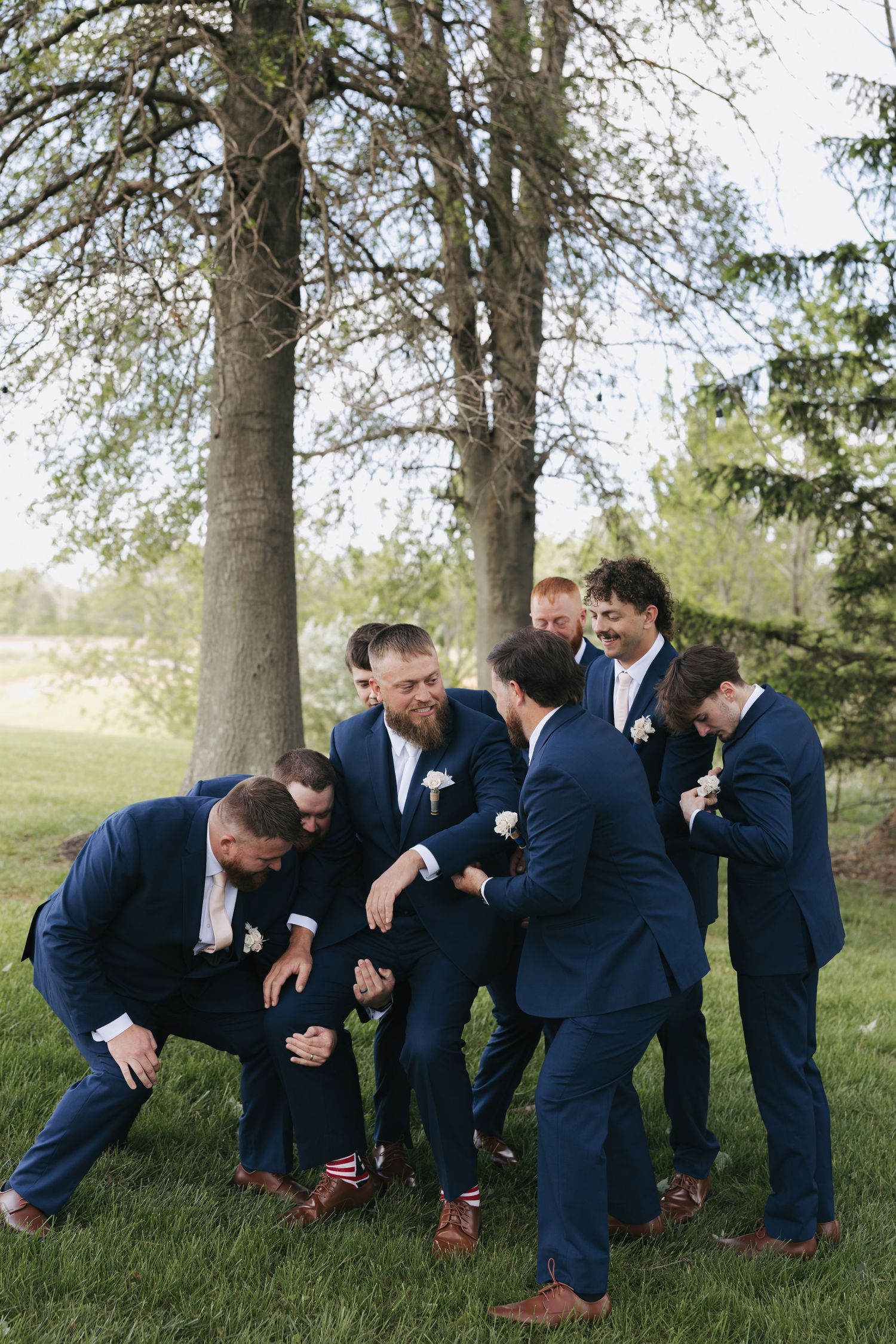 Groomsmen in navy suits playfully roughhousing outdoors near trees before a wedding ceremony.