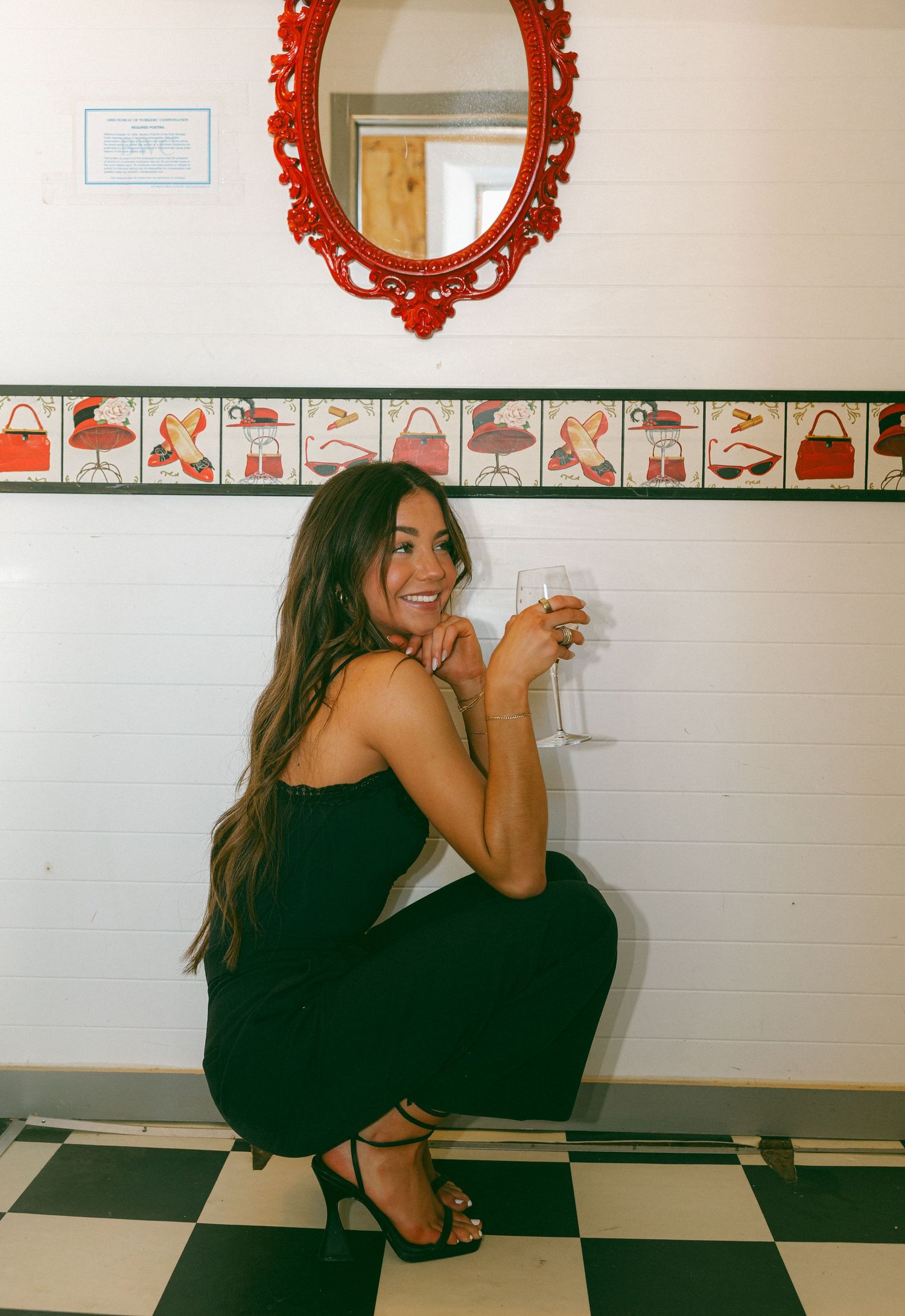 Woman in black dress crouching with wine glass on black and white checkered floor near decorative wall border.