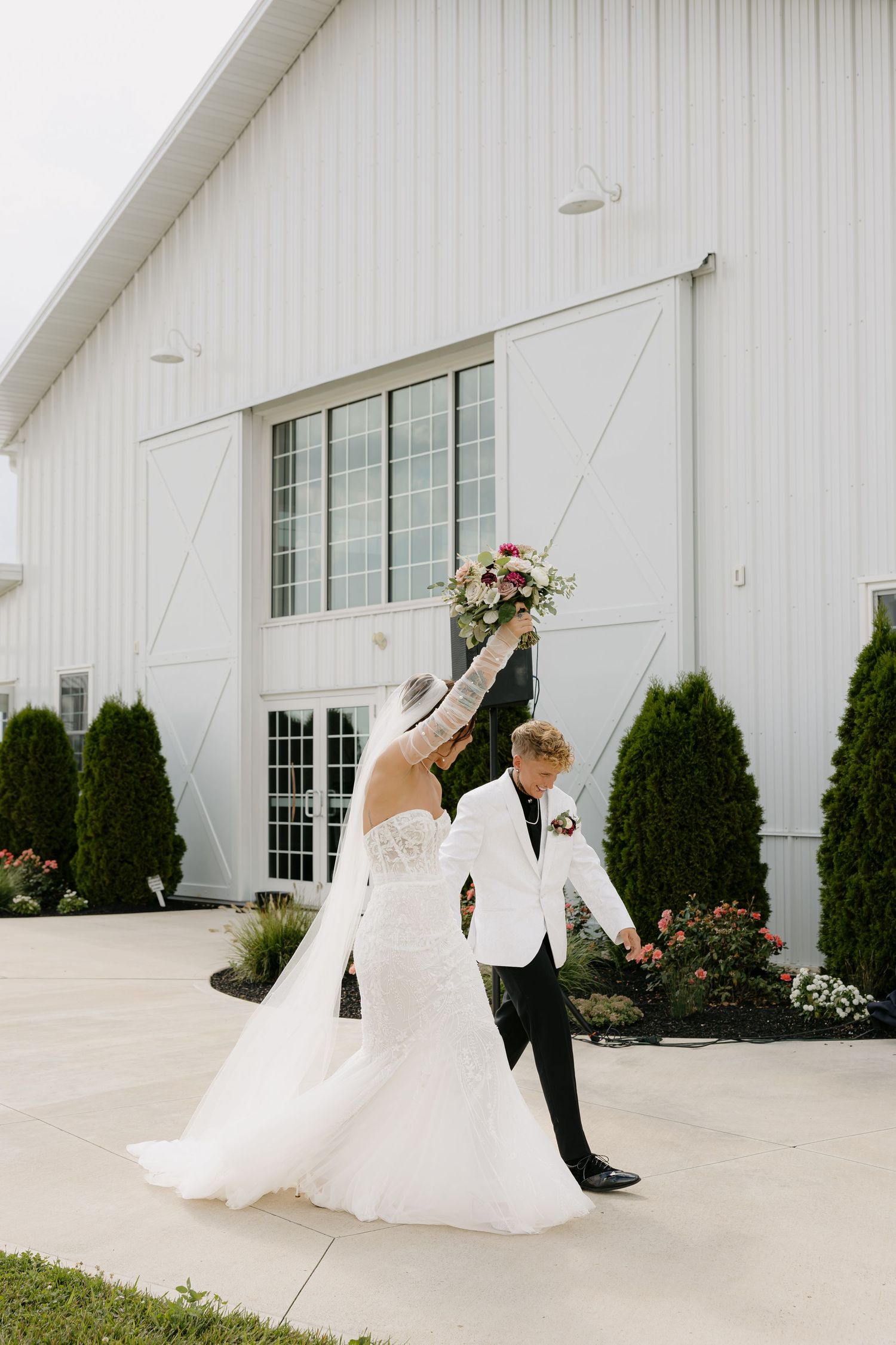 Bride and groom dancing outside white barn wedding venue with bouquet.