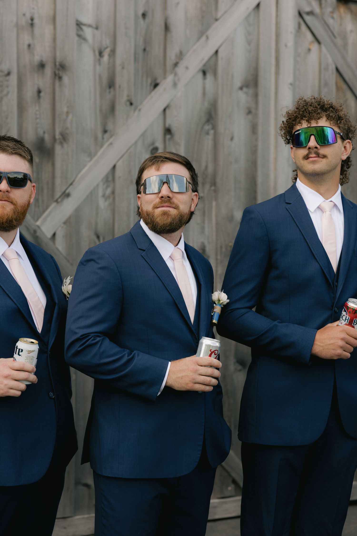 Three groomsmen in navy suits wearing sporty sunglasses holding beer cans by barn.