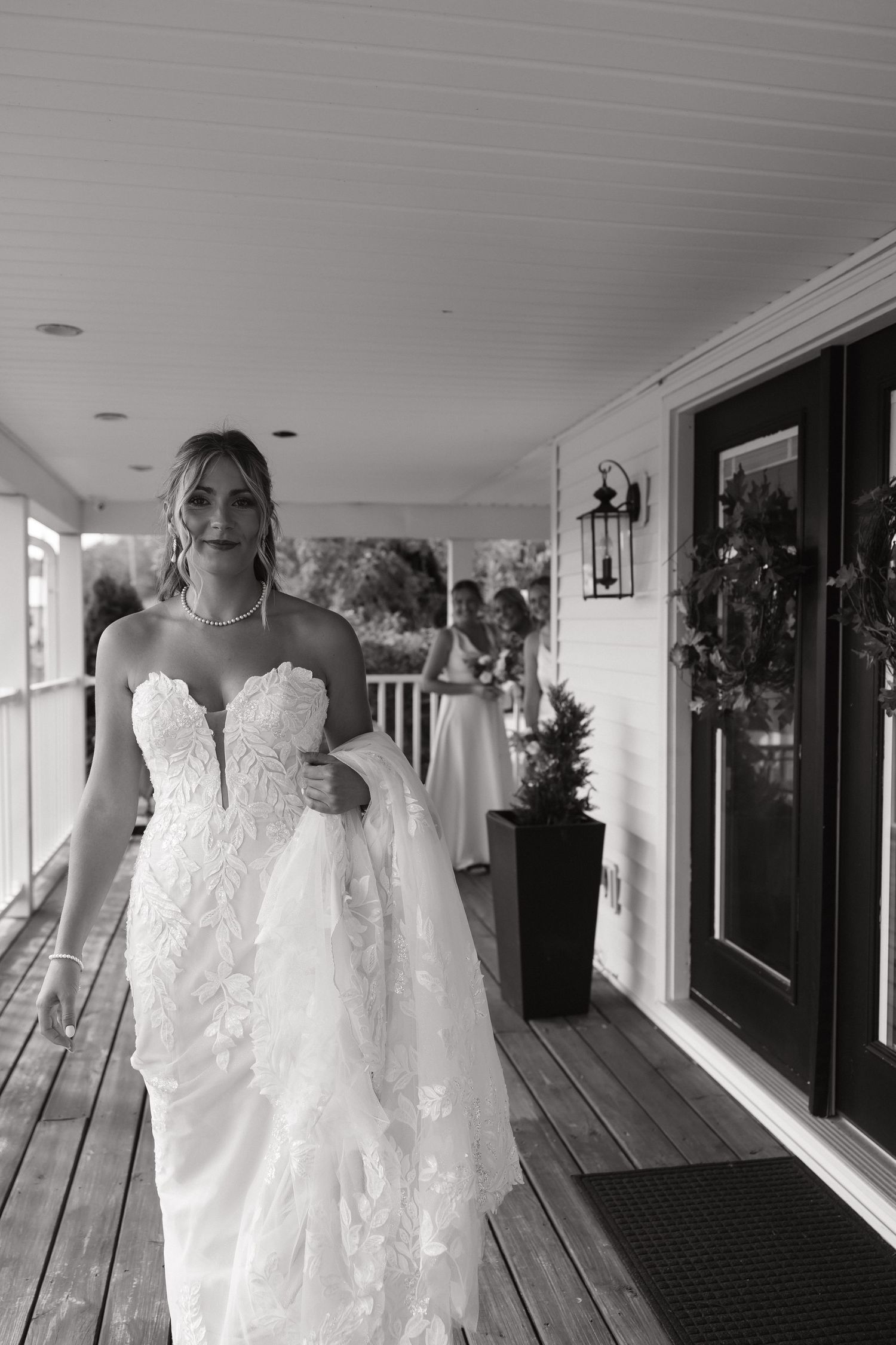 Elegant bride in strapless lace gown walking on a porch in a black and white photo.