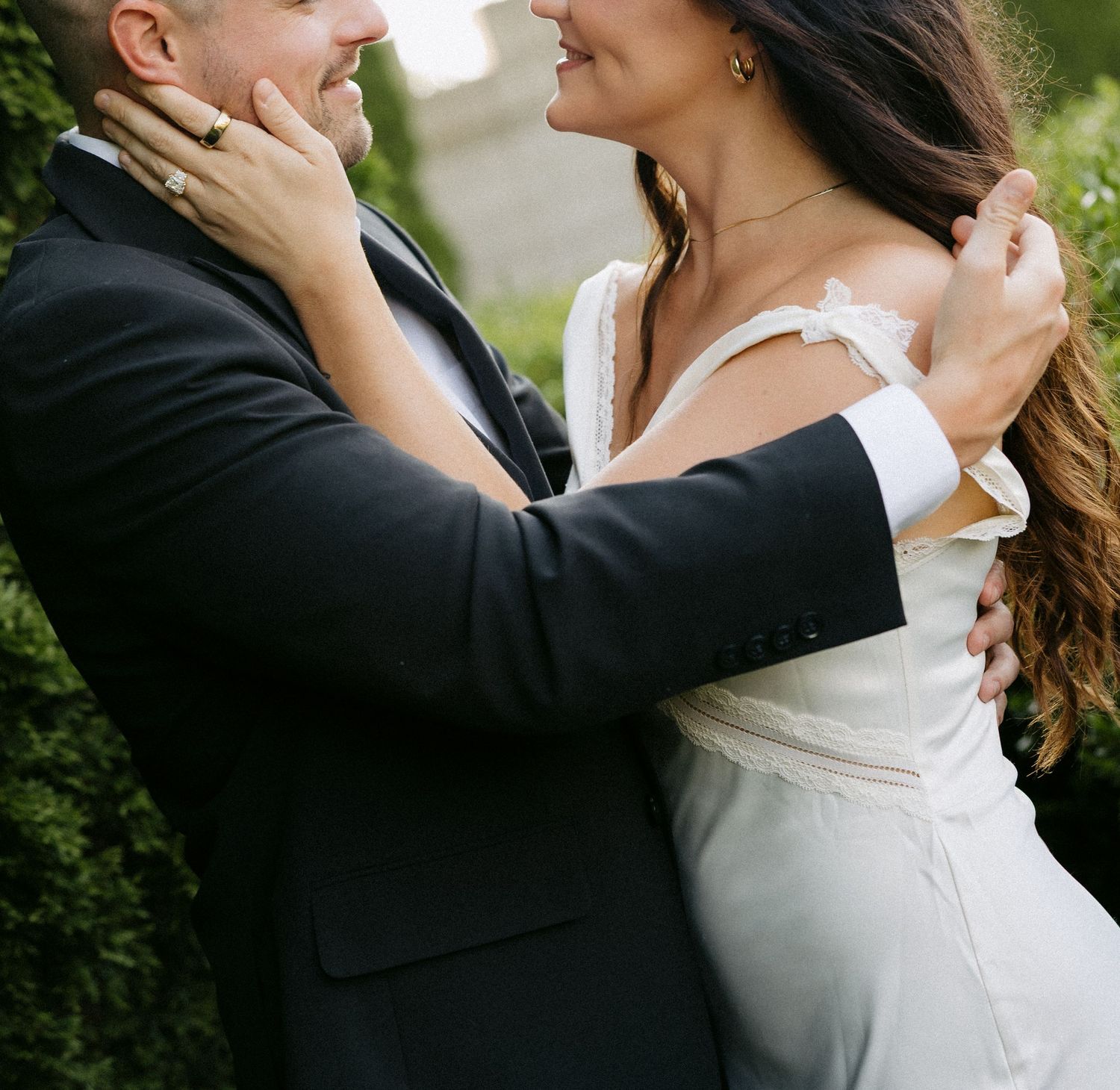 A bride and groom share a romantic embrace outdoors, dressed in wedding attire, surrounded by lush greenery.