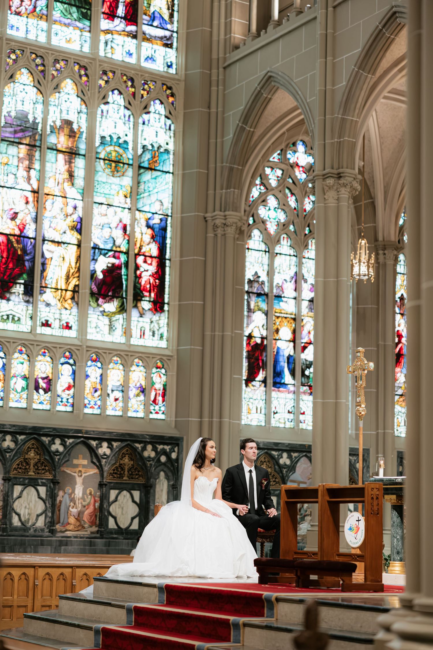 Bride and groom sit together at the altar of a stunning Gothic cathedral with colorful stained glass windows.