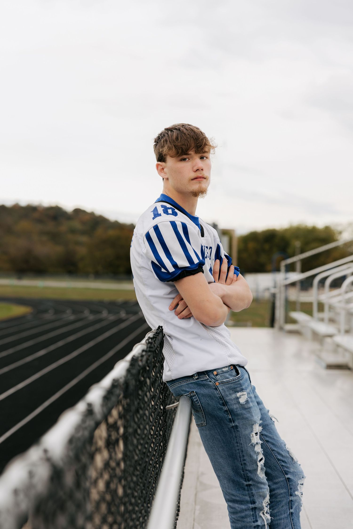 Teen boy in Manchester football jersey and ripped jeans leaning on stadium fence for senior portrait.