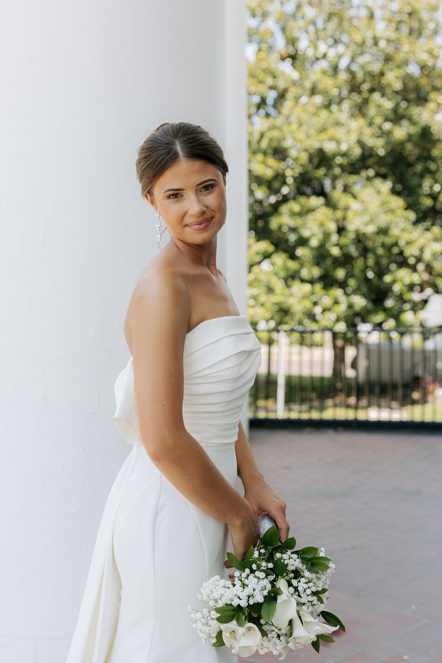 Elegant bride in strapless white gown smiling while holding small white floral bouquet outdoors by white column.