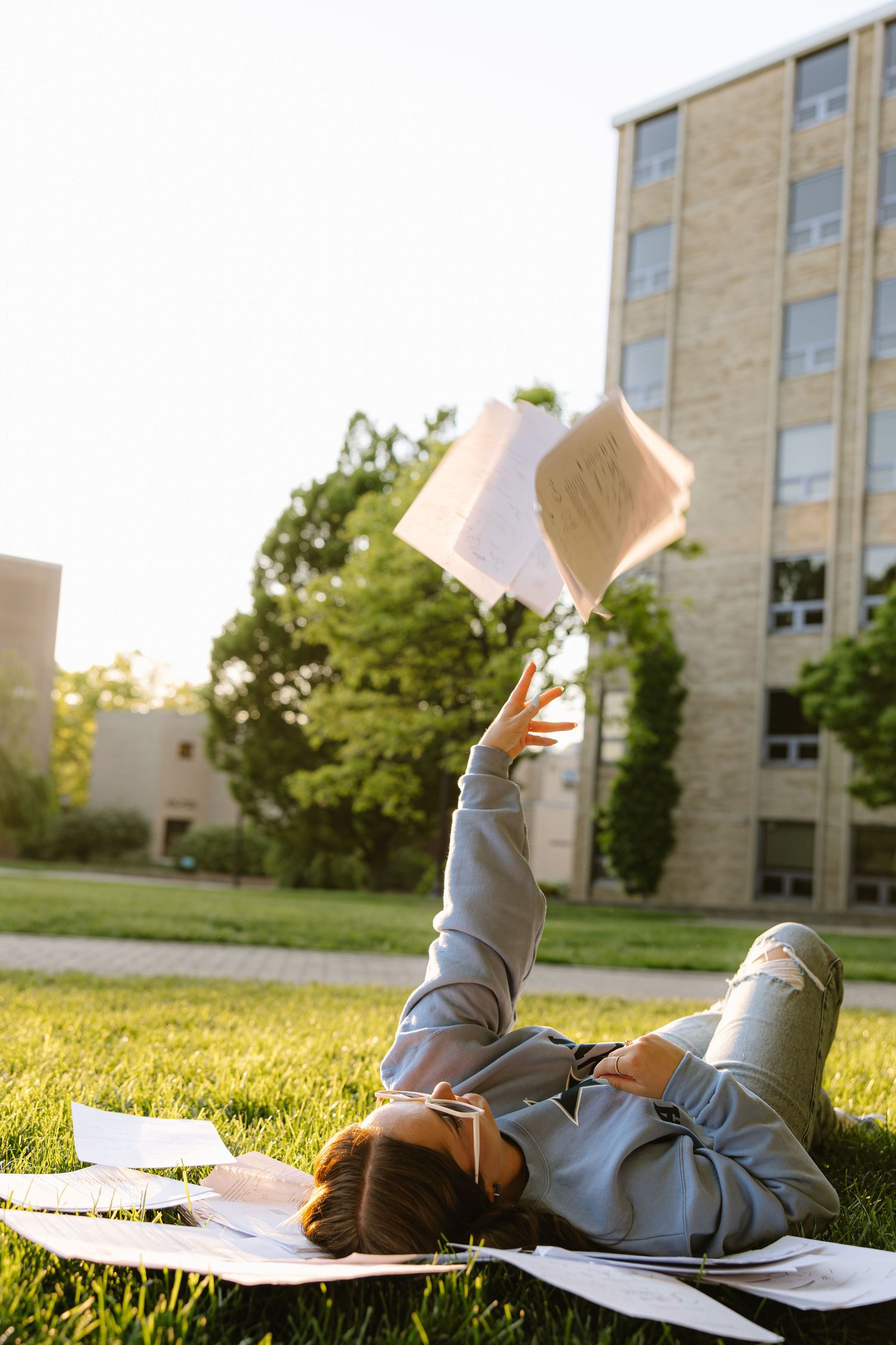 College student lying on grass tossing textbook into the air surrounded by papers on sunny campus lawn.
