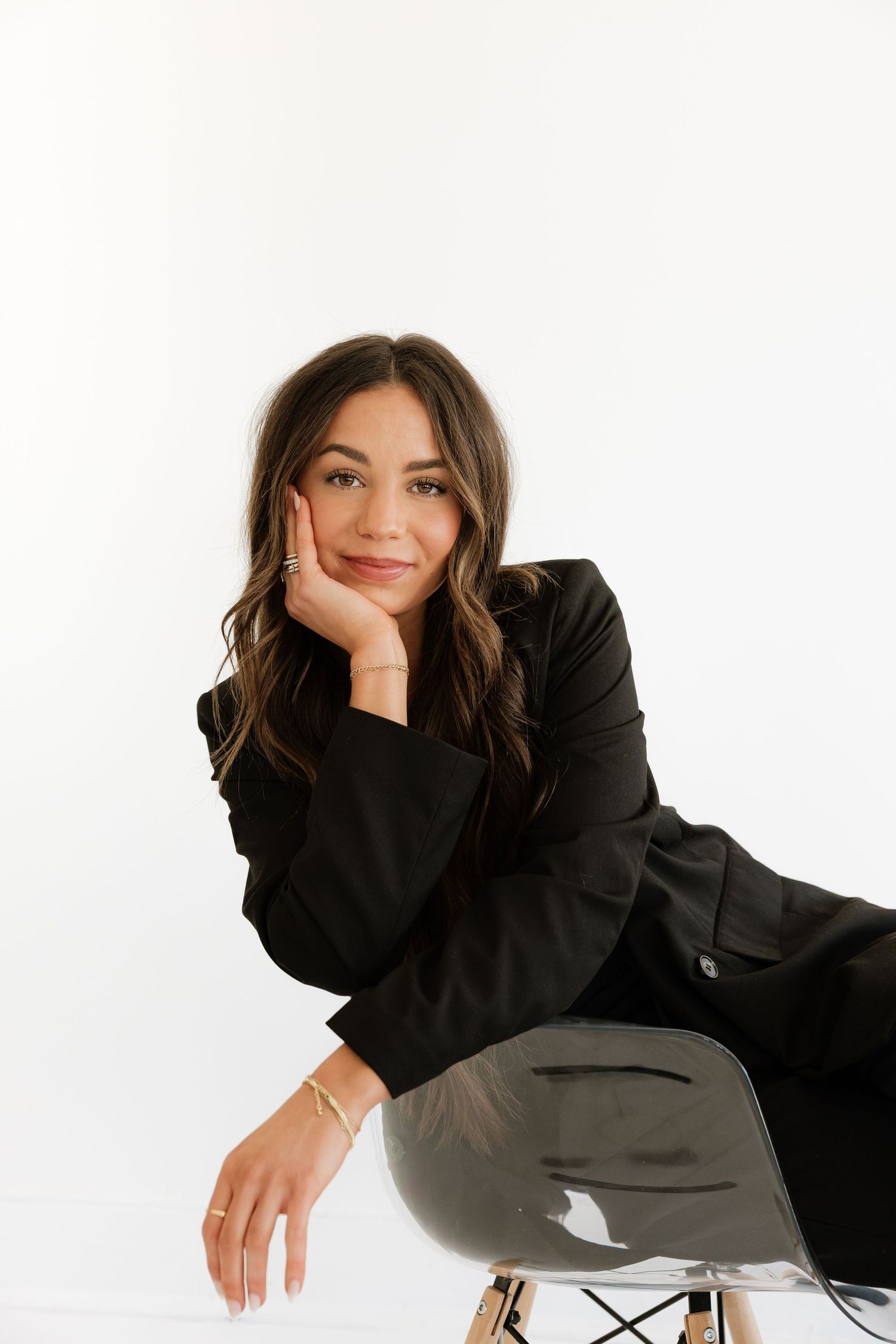 Confident young brunette woman in black blazer posing on clear chair against white studio background.
