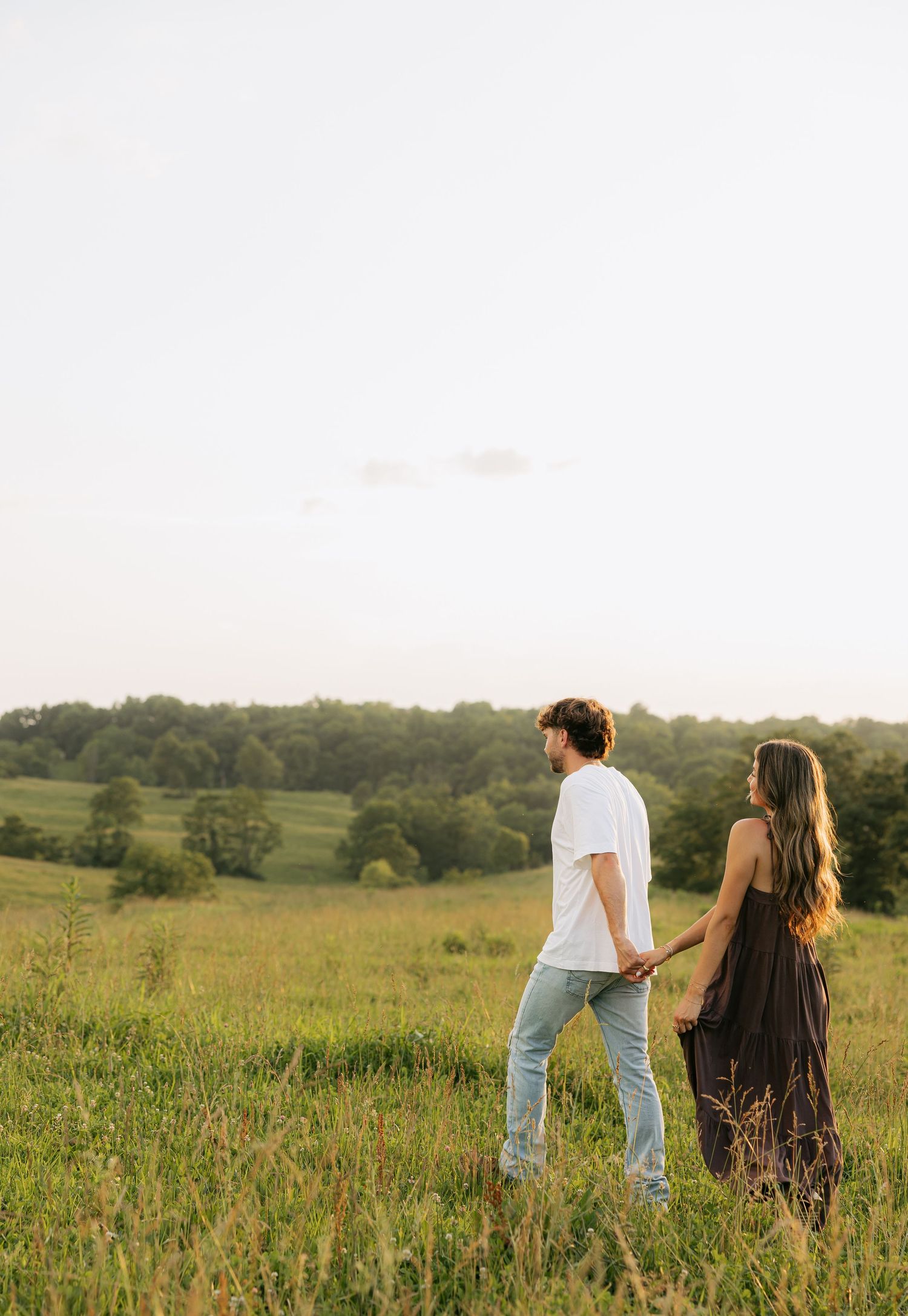 Couple holding hands in a green meadow at golden hour, man in white tee and jeans, woman in black maxi dress.
