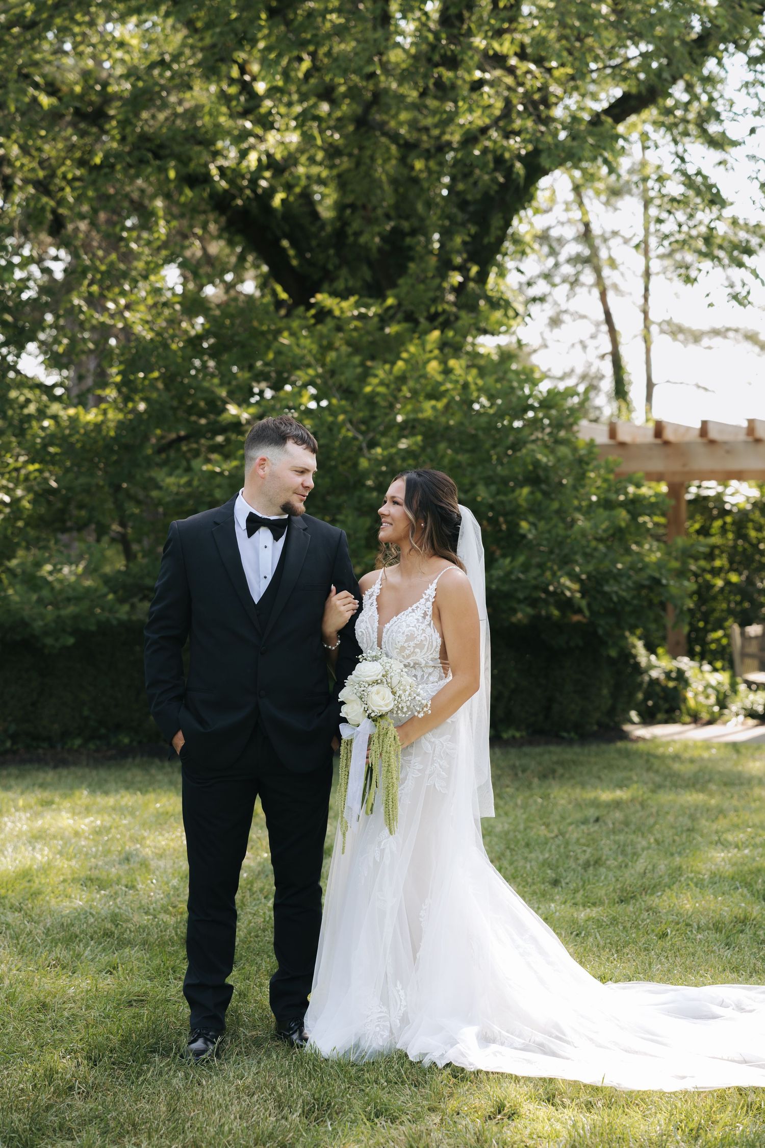 Bride and groom smile at each other in lush garden holding white floral bouquet.