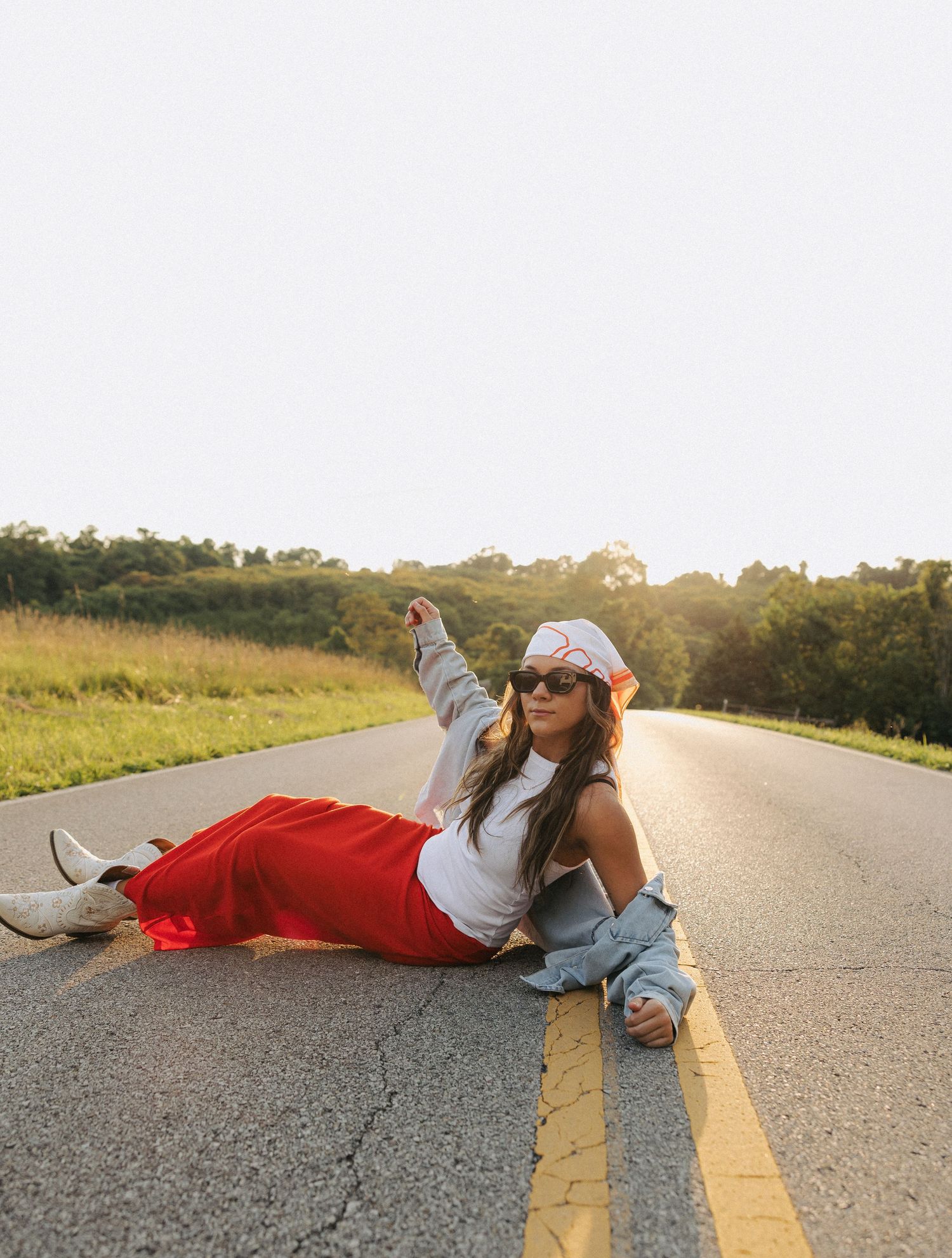 Young woman in red skirt and denim jacket posing on a rural road at golden hour sunset.