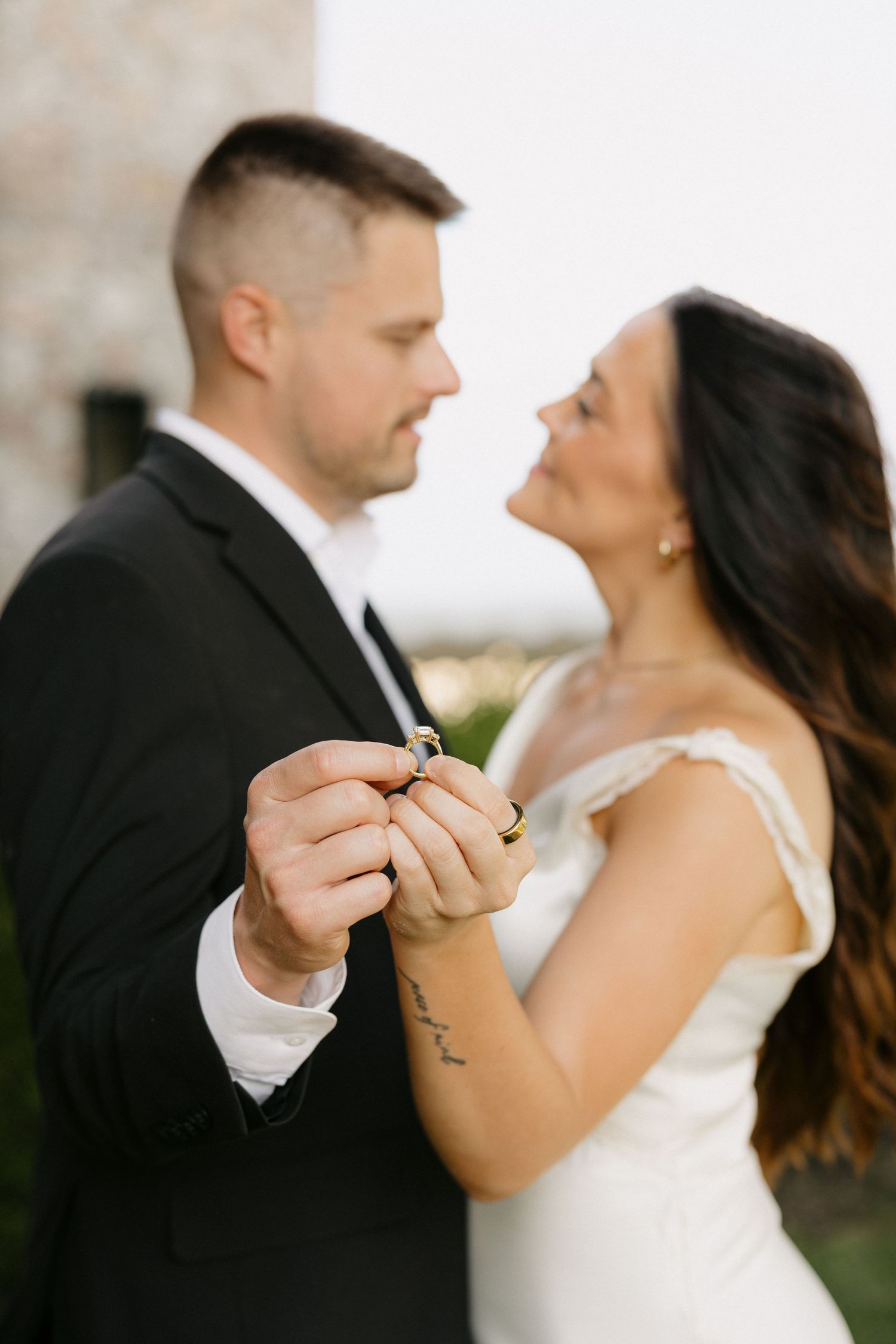 Bride and groom holding wedding rings together, smiling at each other, dressed in white gown and black tuxedo.