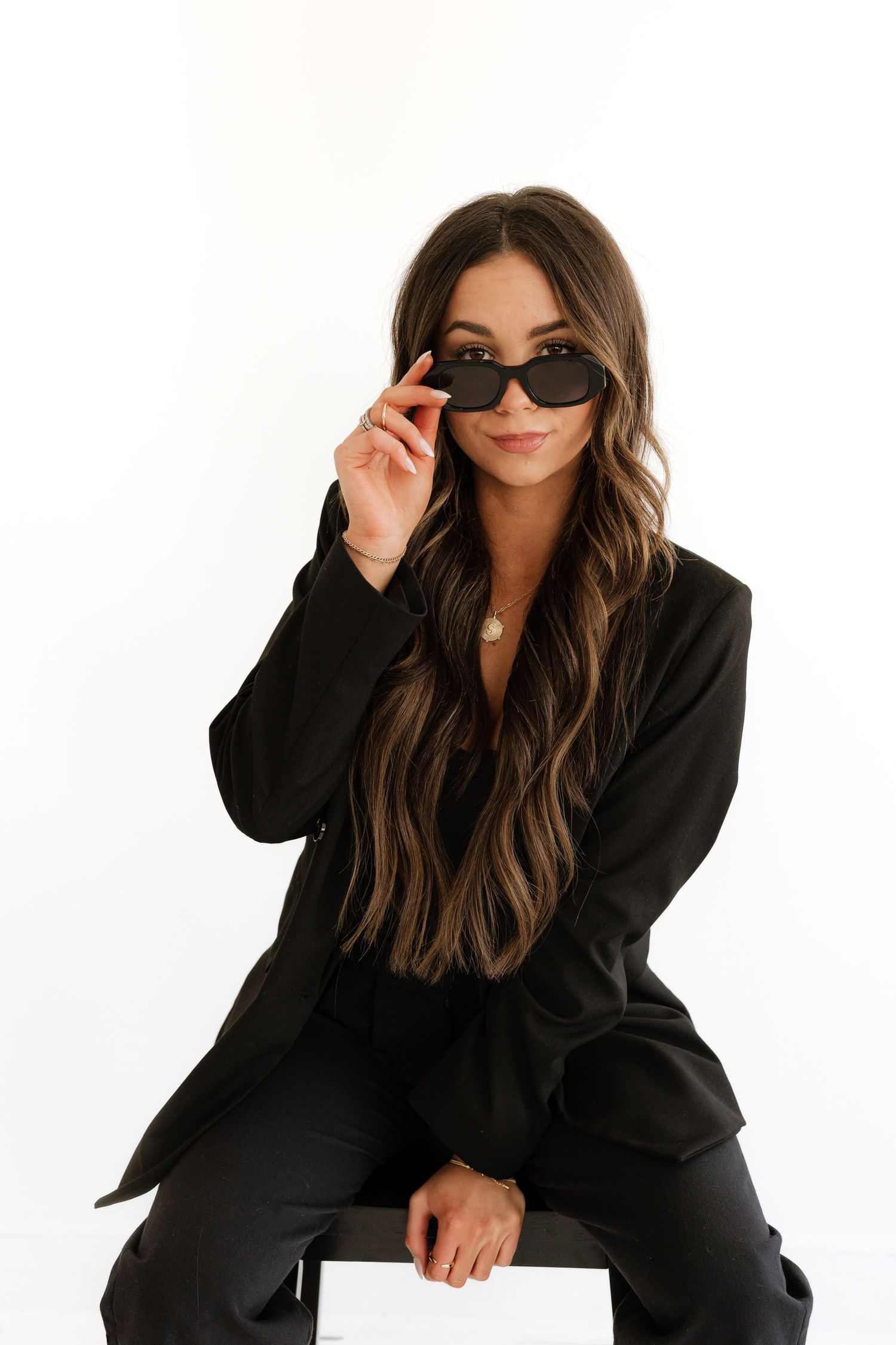 Woman in black blazer and pants sitting confidently, lowering dark sunglasses, with long wavy highlighted hair.