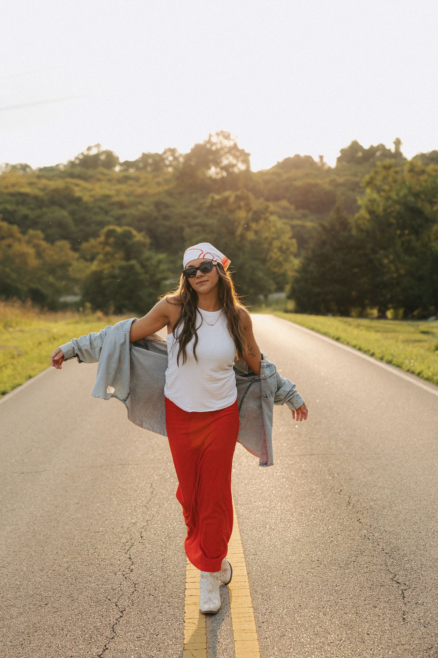 Woman in red pants and denim jacket posing confidently on a tree-lined road at sunset.