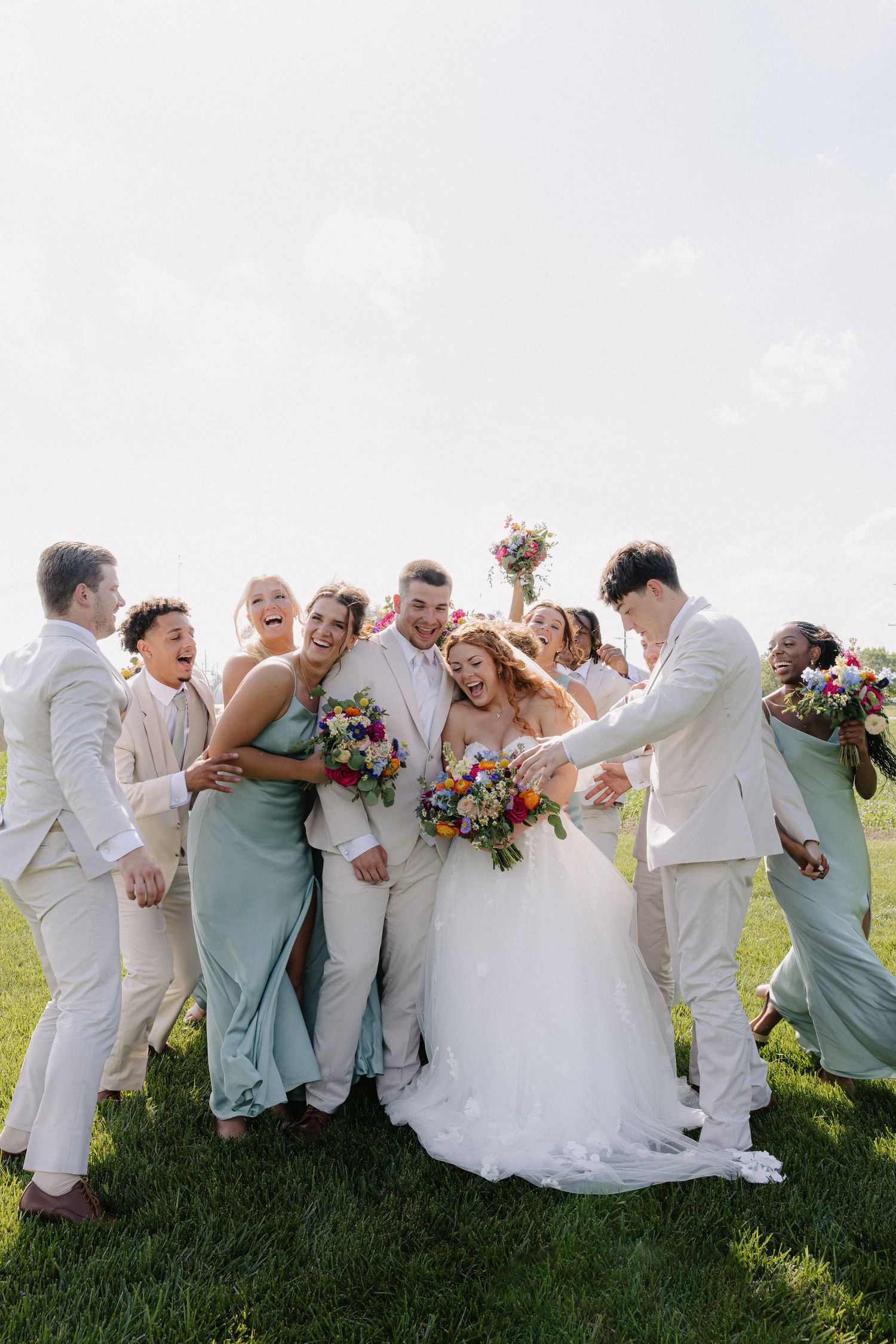 Joyful wedding party celebrates on green lawn with bride, groom, bridesmaids in sage and groomsmen.