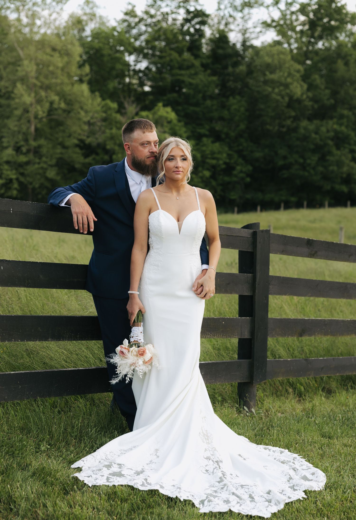 Bride in fitted white gown with lace train holds bouquet beside groom in navy suit at rustic wooden fence.