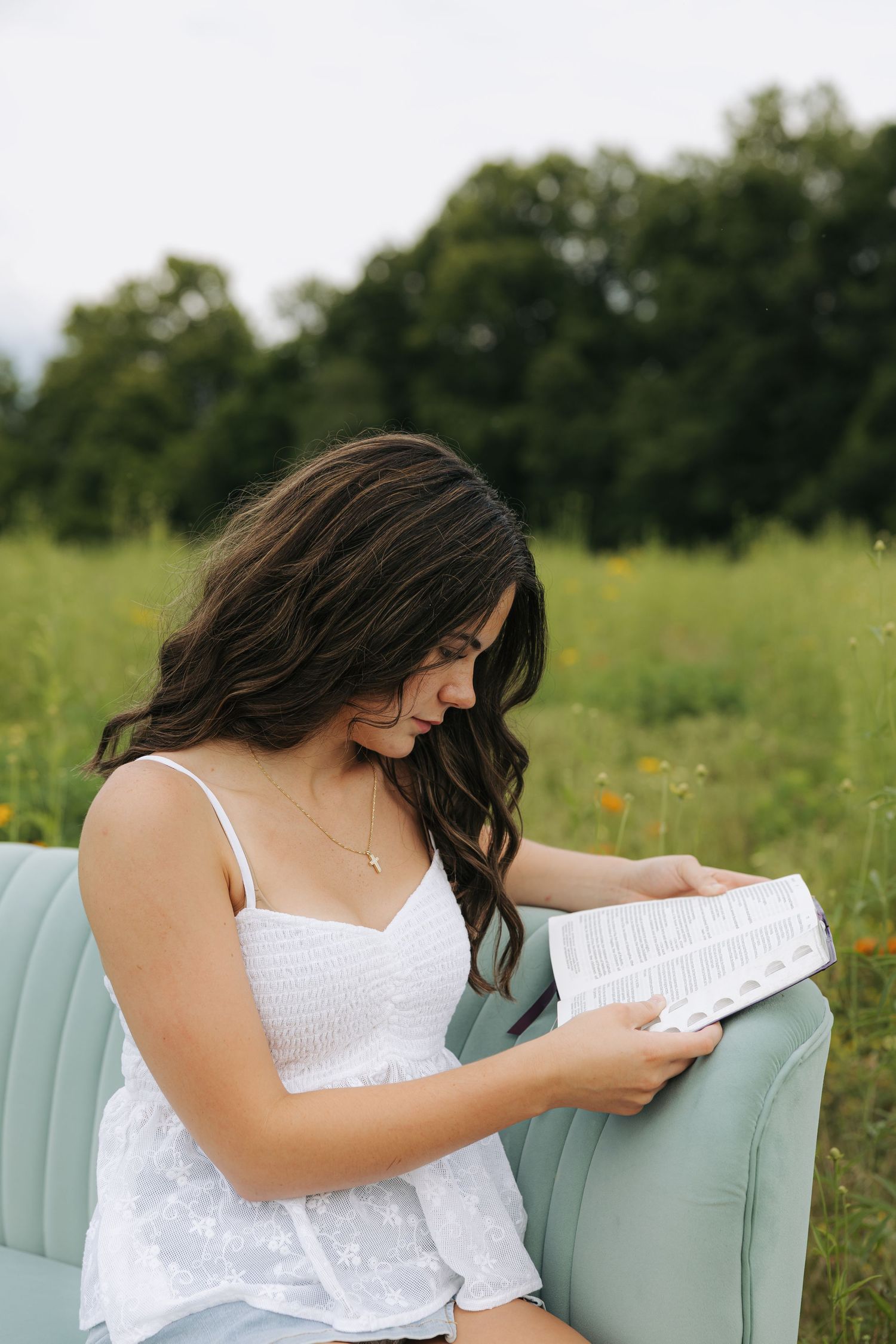 Young brunette woman reading a book while sitting on a mint sofa in a green field.