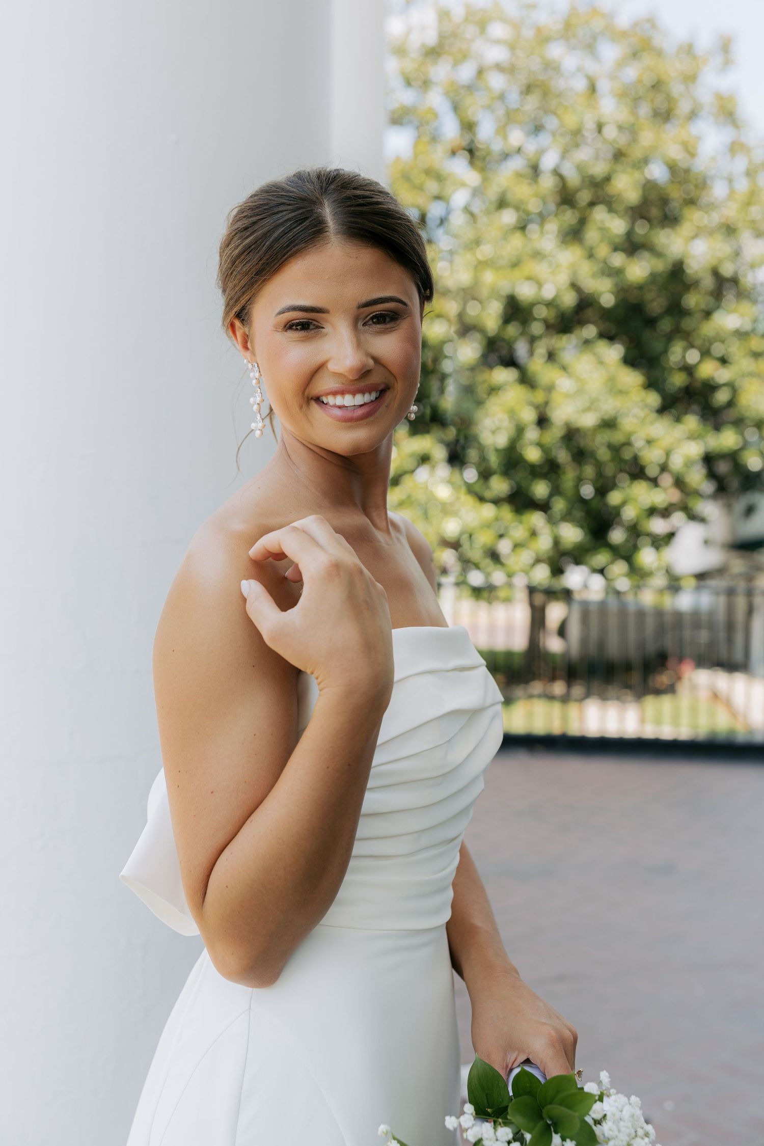 Radiant bride in strapless white dress smiling brightly while standing by white pillar with lush trees behind.