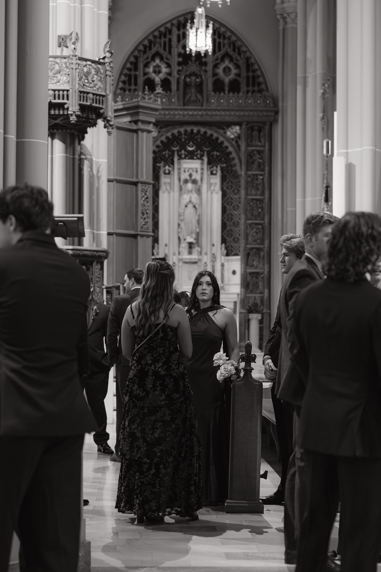 Black and white photo of elegantly dressed wedding guests socializing inside a grand cathedral with ornate stone arches.