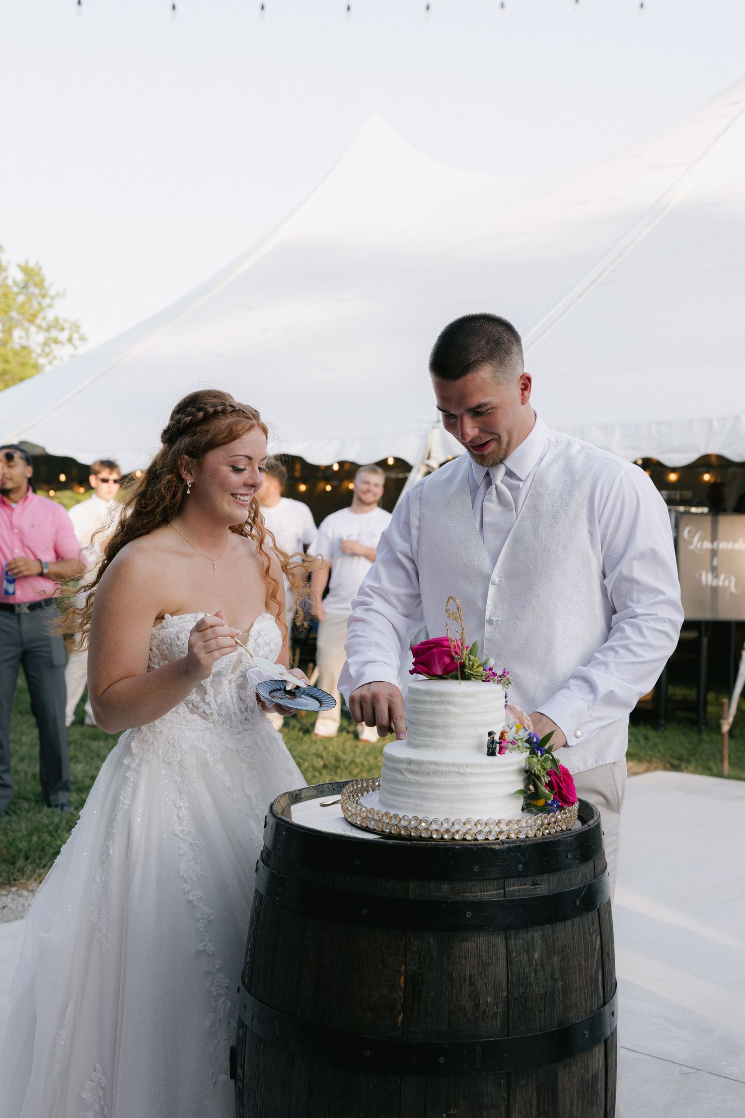 Bride and groom cut tiered white wedding cake displayed on rustic barrel at reception.