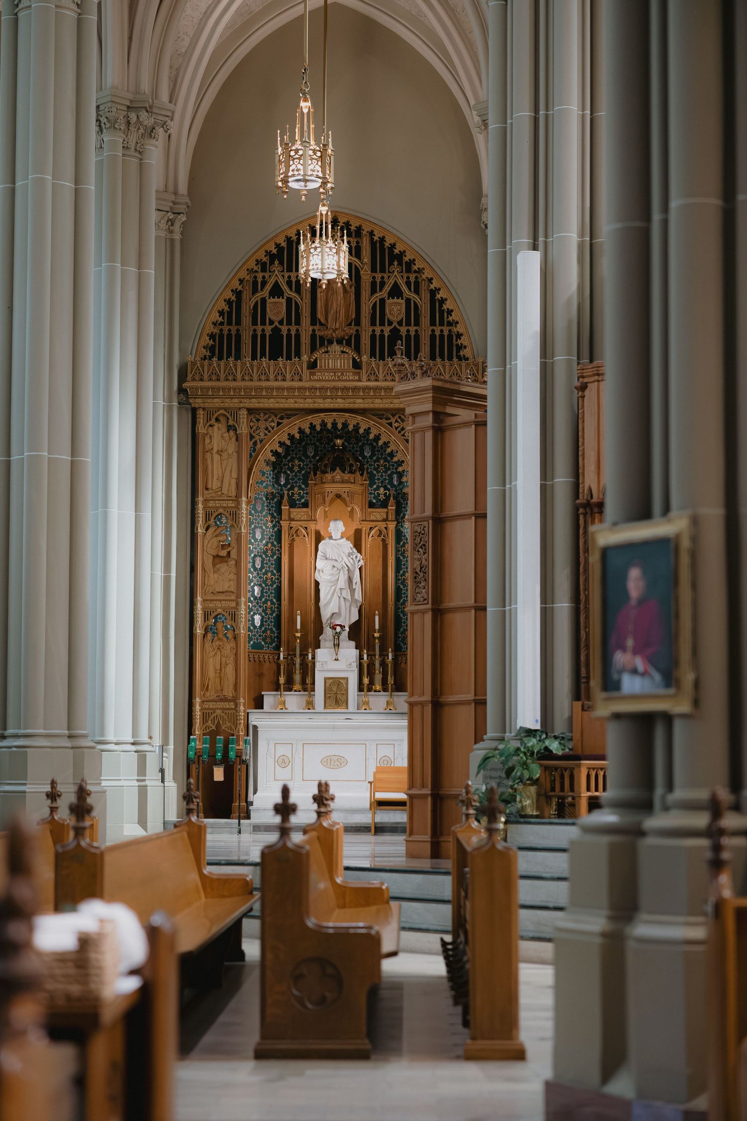 Grand cathedral interior featuring Gothic arches, wooden pews, ornate altar, and a white statue illuminated by chandelier light.
