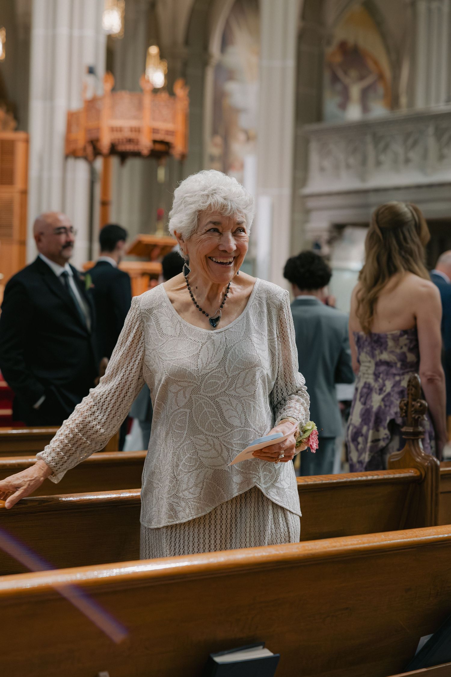 Smiling elderly woman in cream lace outfit standing between church pews at a wedding ceremony inside a grand cathedral.