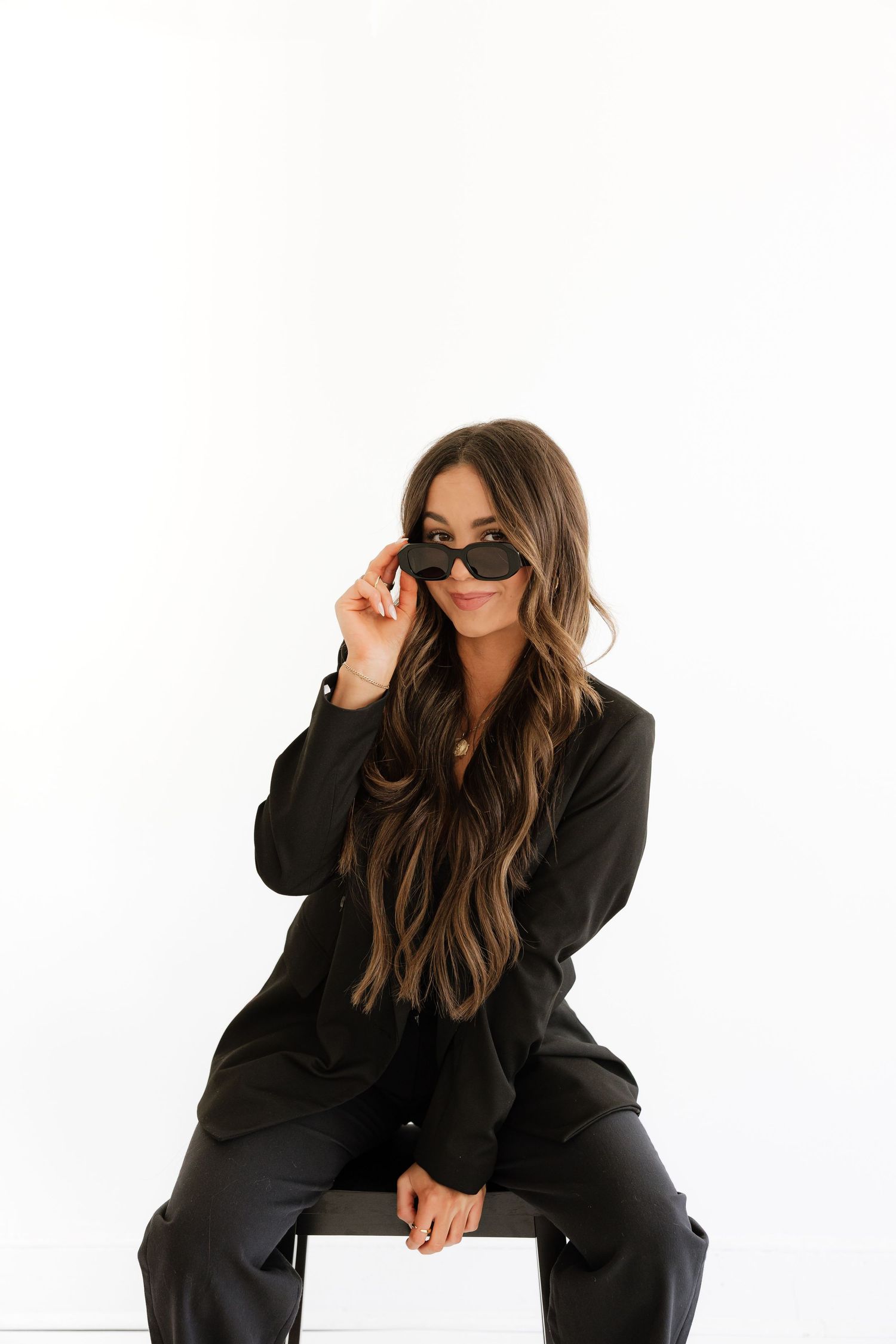 Brunette woman in black suit sitting on stool, lowering sunglasses, white background.