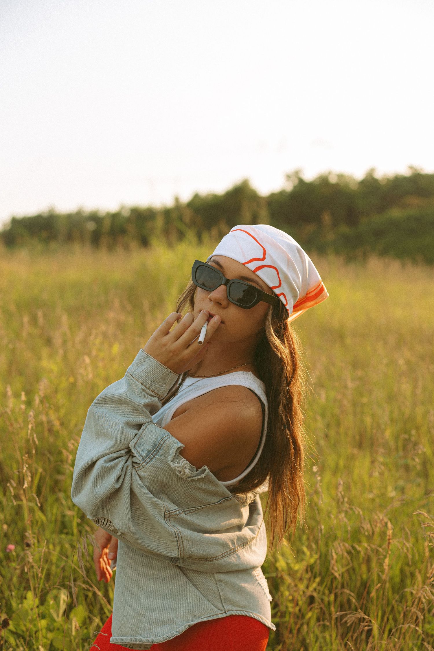 Stylish woman in sunglasses, white bandana, and denim jacket smoking in a golden sunlit grassy field.