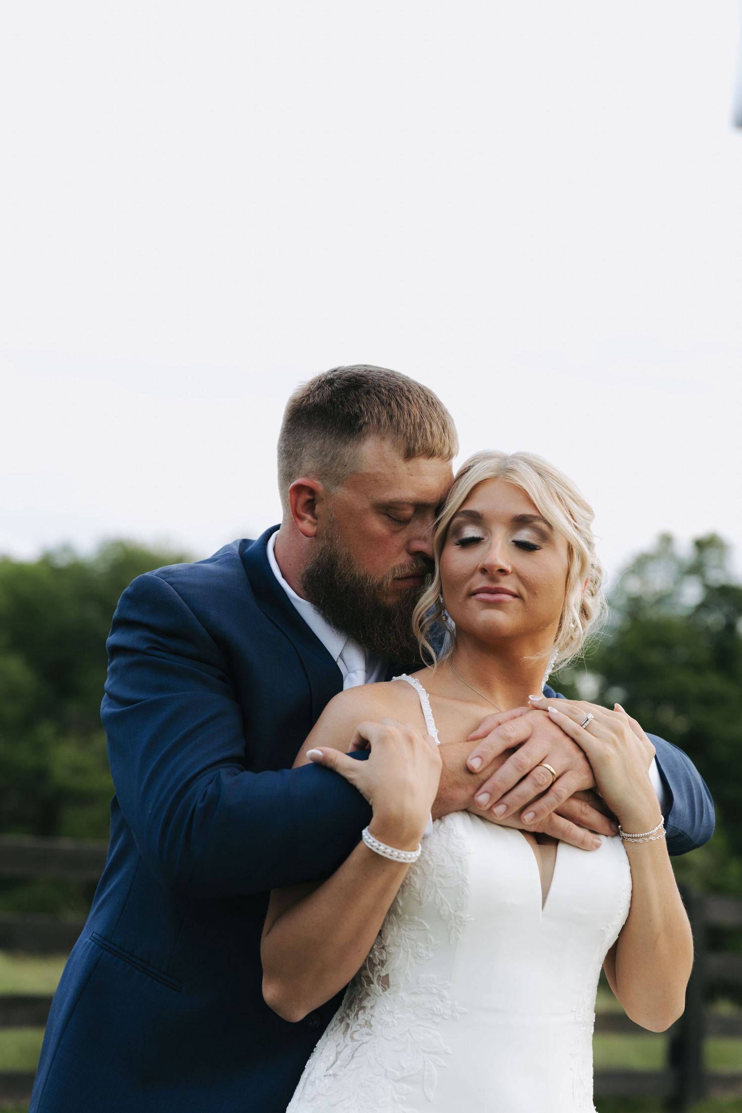 Bearded groom in navy suit kisses blonde bride tenderly on cheek outdoors.