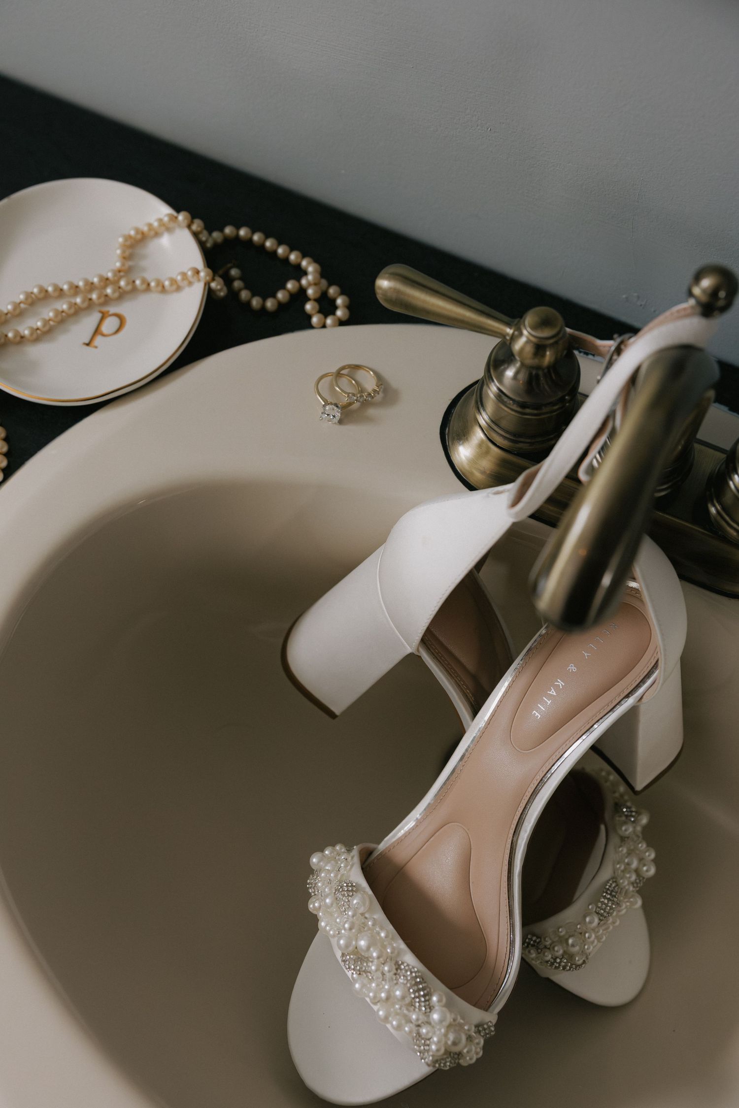 White bridal heels with pearl details displayed in a vintage sink with jewelry and rings.