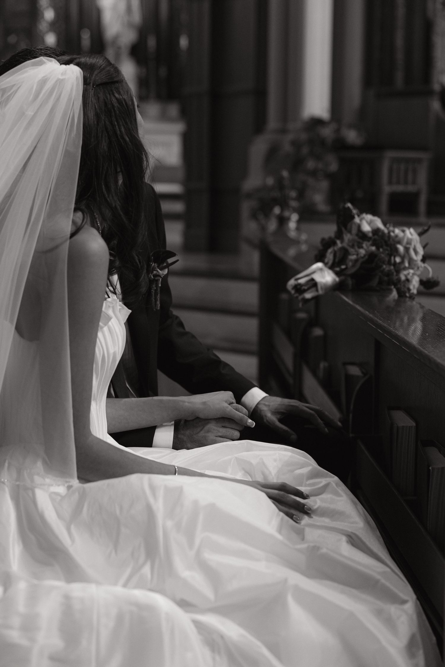 Black and white photo of bride and groom holding hands on church pew, bride in white wedding dress.