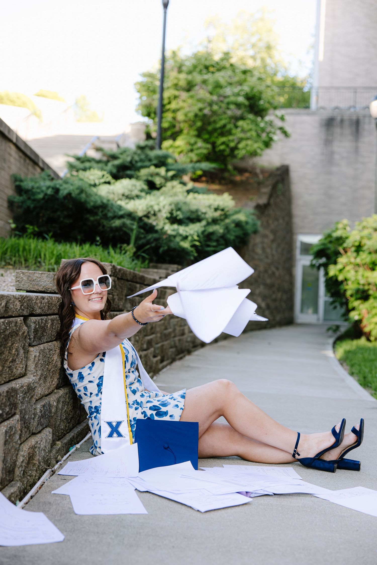 Smiling graduate in floral dress and white sunglasses sitting outdoors tossing papers with blue diploma folder.