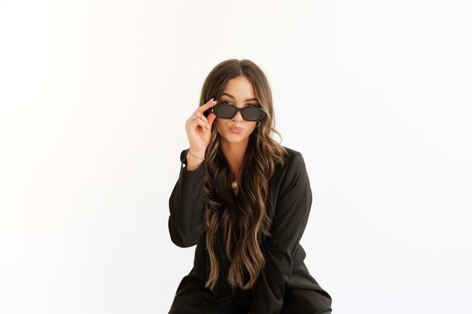 Confident brunette woman in black blazer adjusting sunglasses against white background.