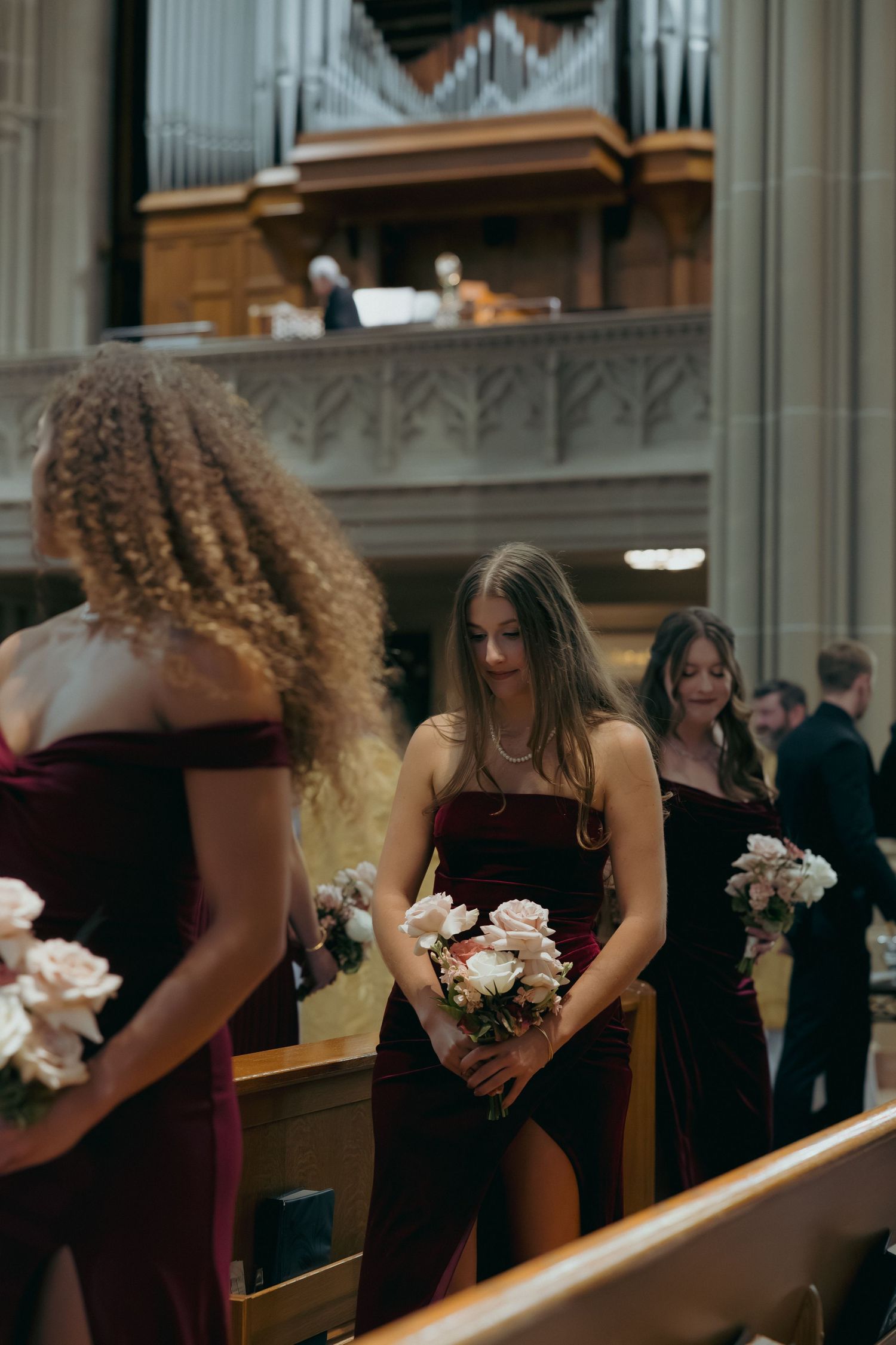 Bridesmaids in burgundy velvet dresses holding floral bouquets inside an elegant church during a wedding ceremony.