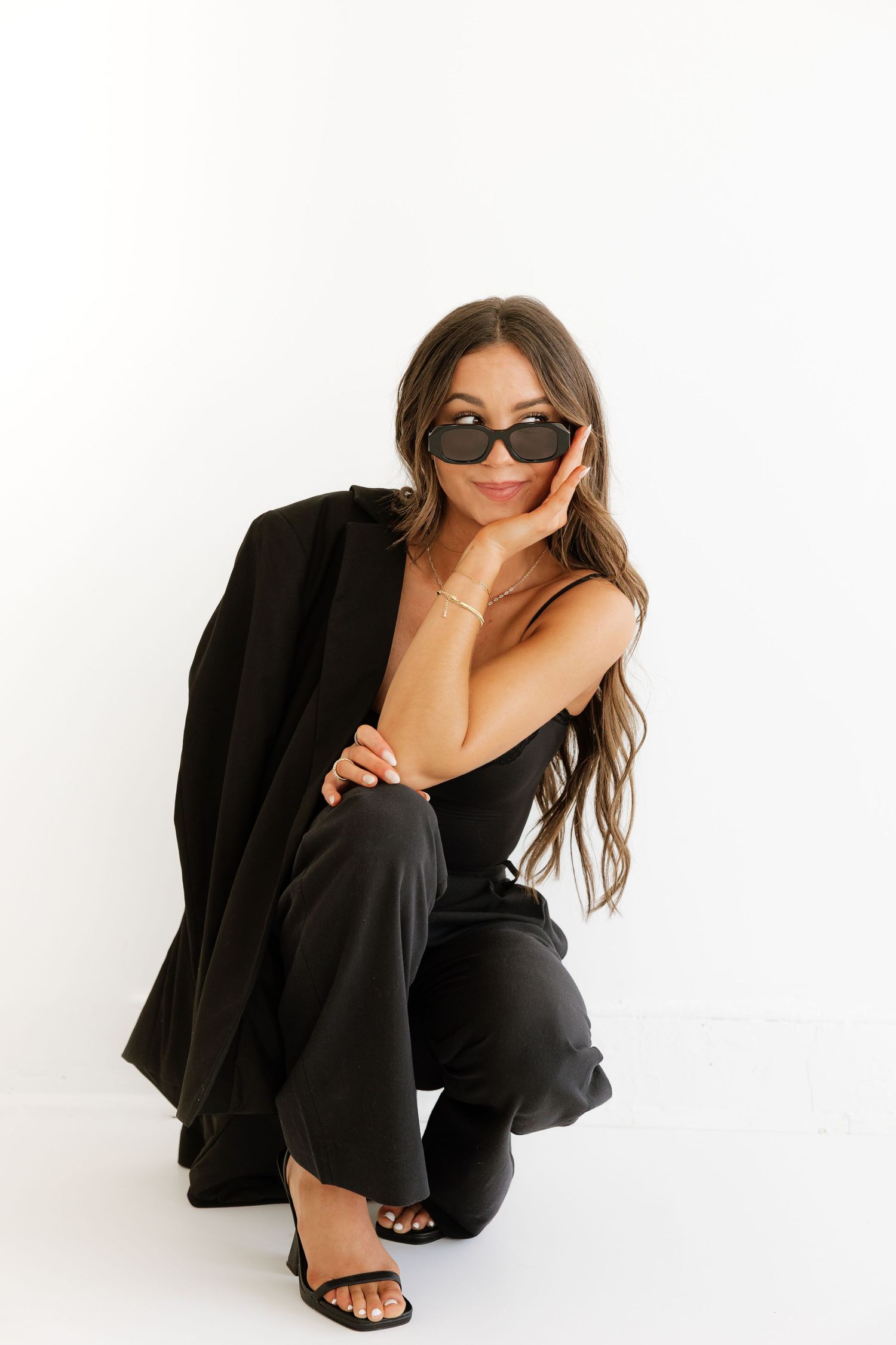 Fashionable brunette woman crouching in all-black outfit and sunglasses against a white studio backdrop.