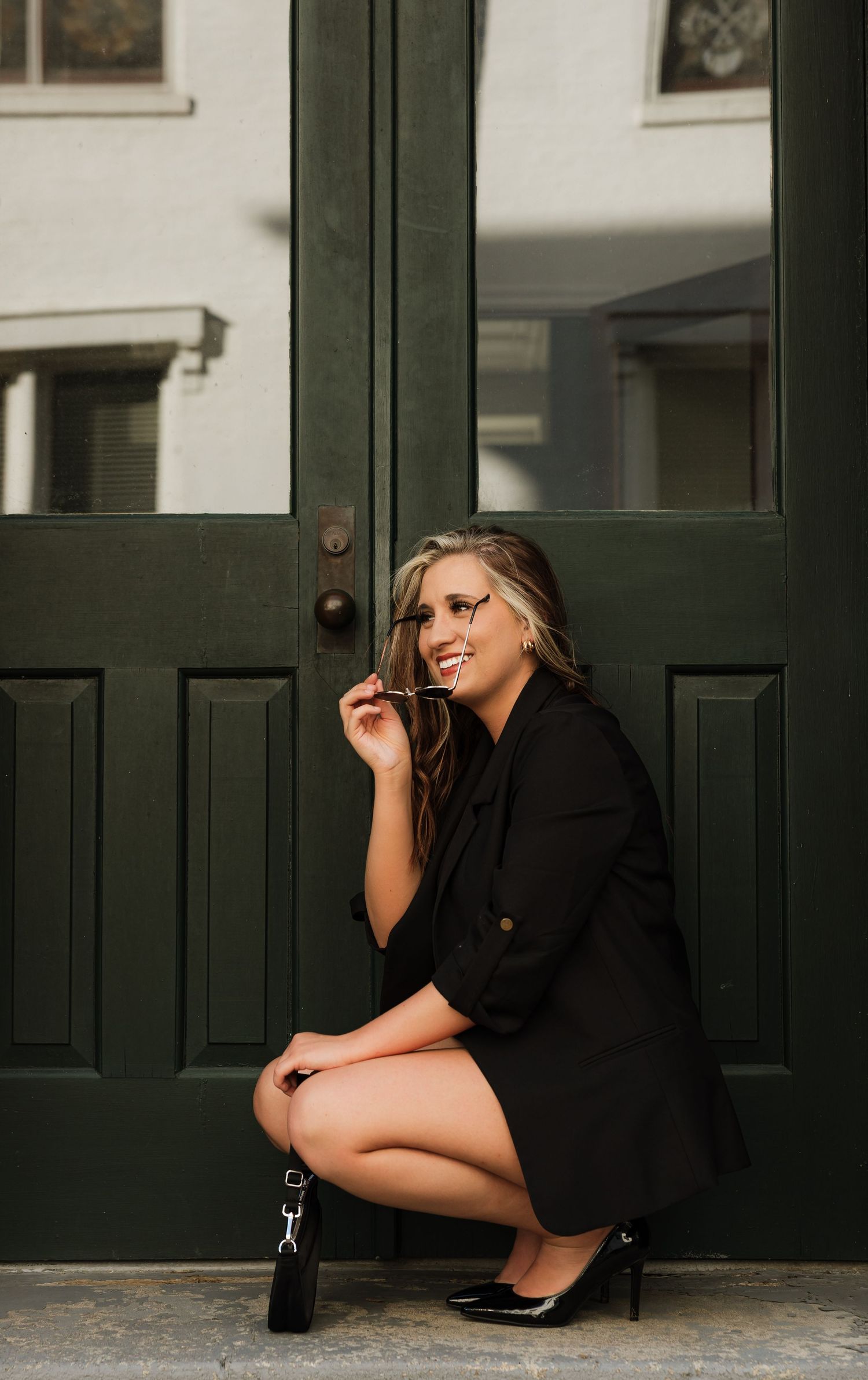 Smiling blonde woman in black blazer dress crouching by dark green doors holding sunglasses and purse.