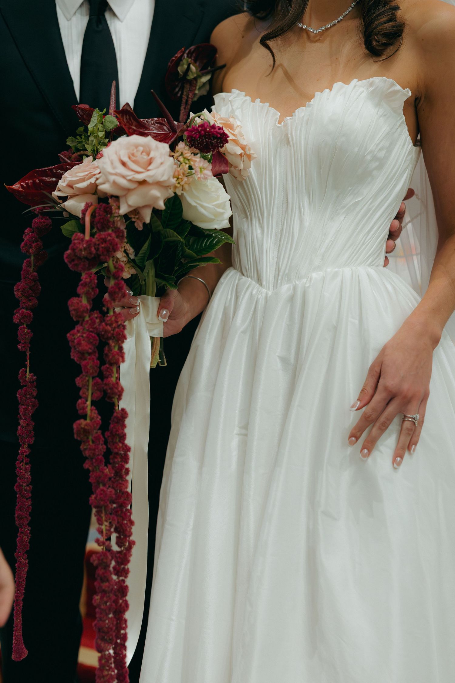 Close-up of bride in strapless white wedding gown holding a cascading bouquet of burgundy and blush roses with ribbons.