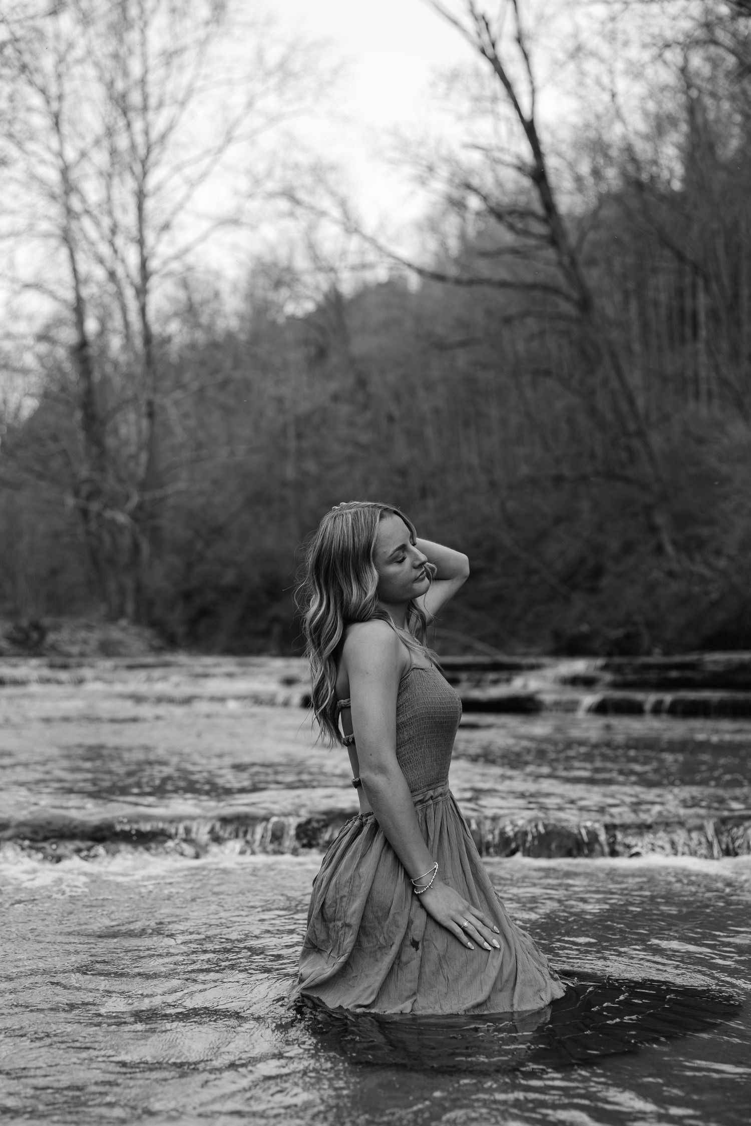 Black and white portrait of a woman in a dress standing in a flowing river surrounded by trees.