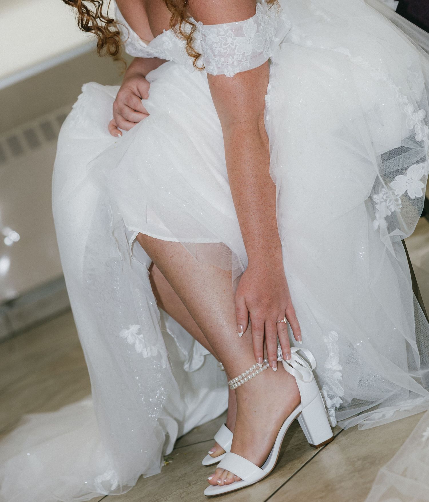 Bride in white lace gown fastening pearl ankle strap on white block heel wedding shoe.