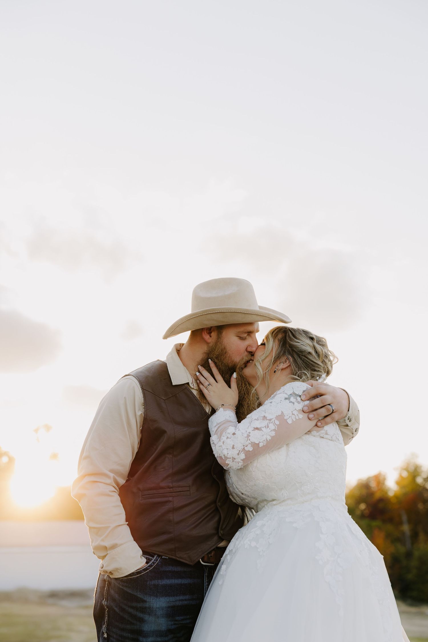 Bride and groom sharing a romantic kiss at sunset, groom wearing a cowboy hat and vest at an outdoor wedding.